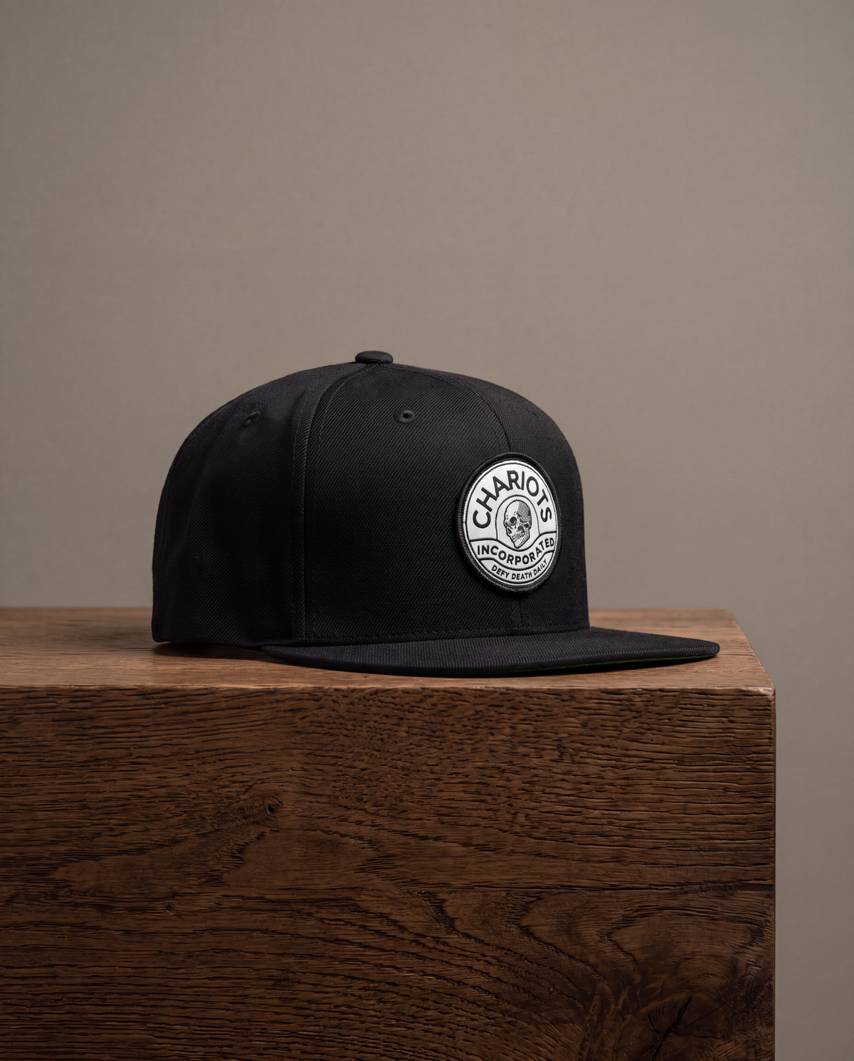 A black flat-brim cap with a circular white and black embroidered patch on the front, displayed on a wooden block against a neutral beige background.