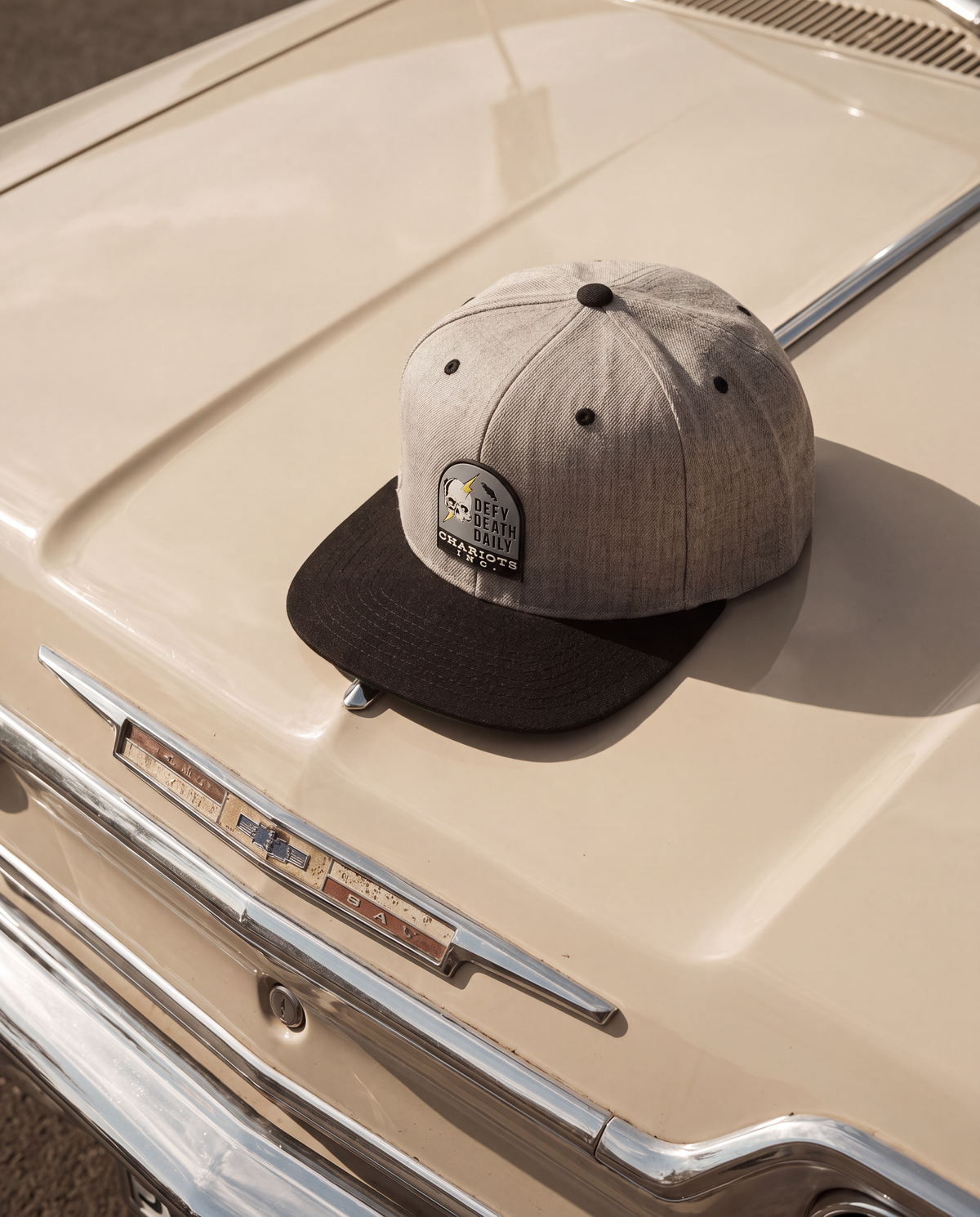 A two-tone snapback cap with a heather gray crown and a dark brown flat brim, resting on the glossy beige hood of a vintage car. The cap features a circular embroidered patch on the front.
