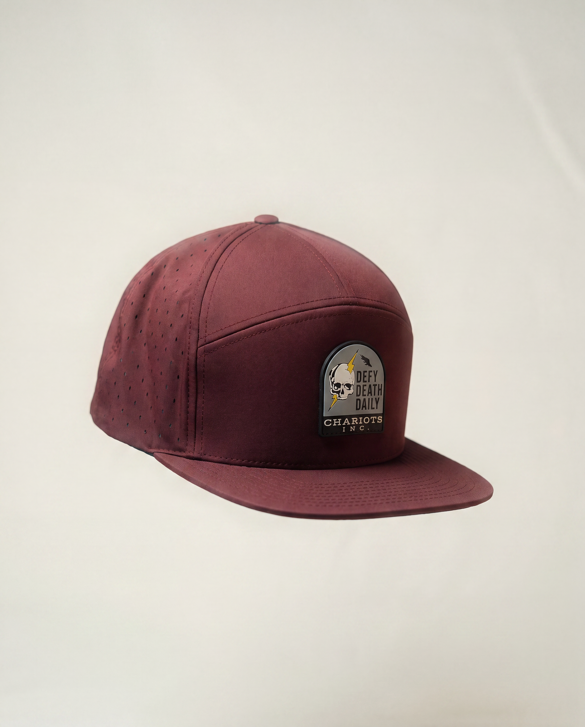 A maroon baseball cap with perforated side panels and a small circular patch on the front, photographed against a plain off-white background.