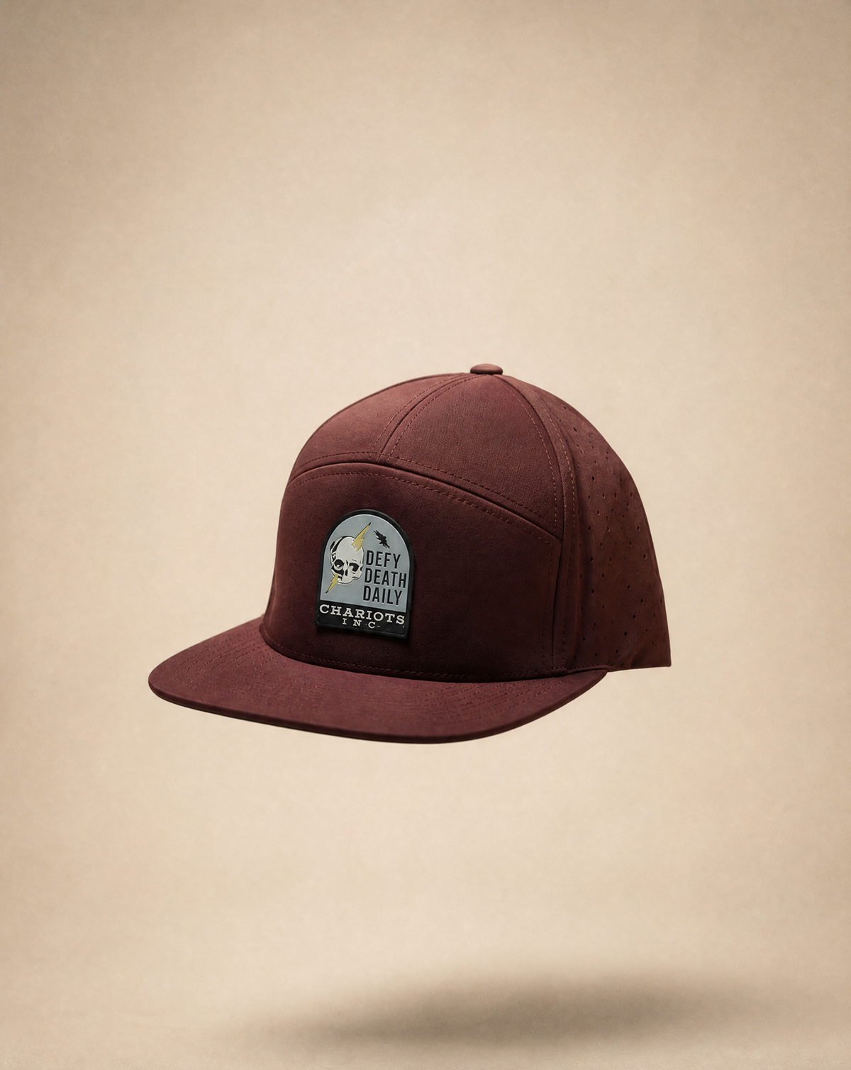 A maroon flat-brim baseball cap with a circular 'DEFY DEATH DAILY CHARIOTS INC' logo patch on the front, shown floating against a textured beige background.