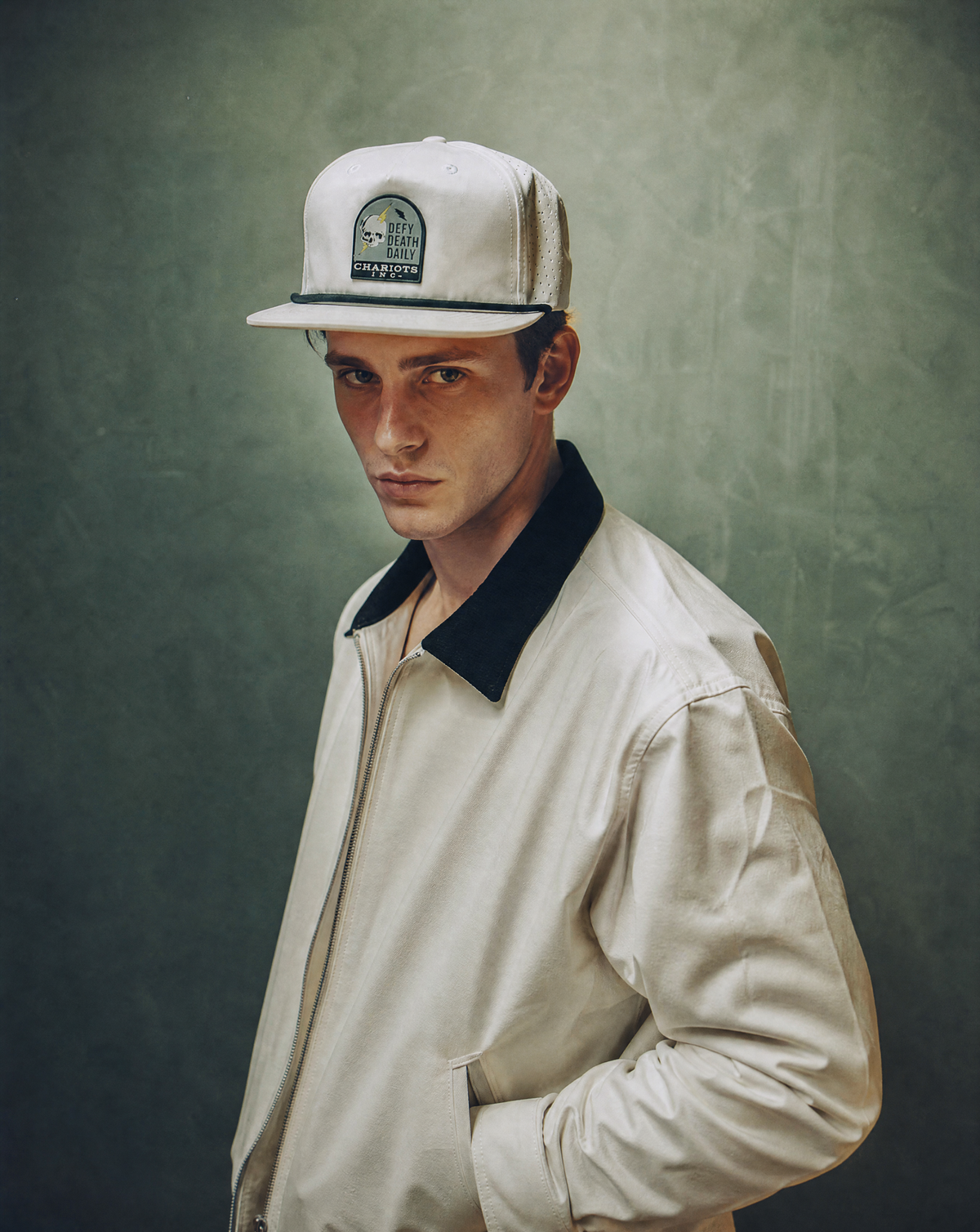 A man wearing a light beige baseball cap with a dark green and white 'Brixton' logo patch, paired with a matching light beige jacket featuring a dark navy collar, posed against a textured dark green background.