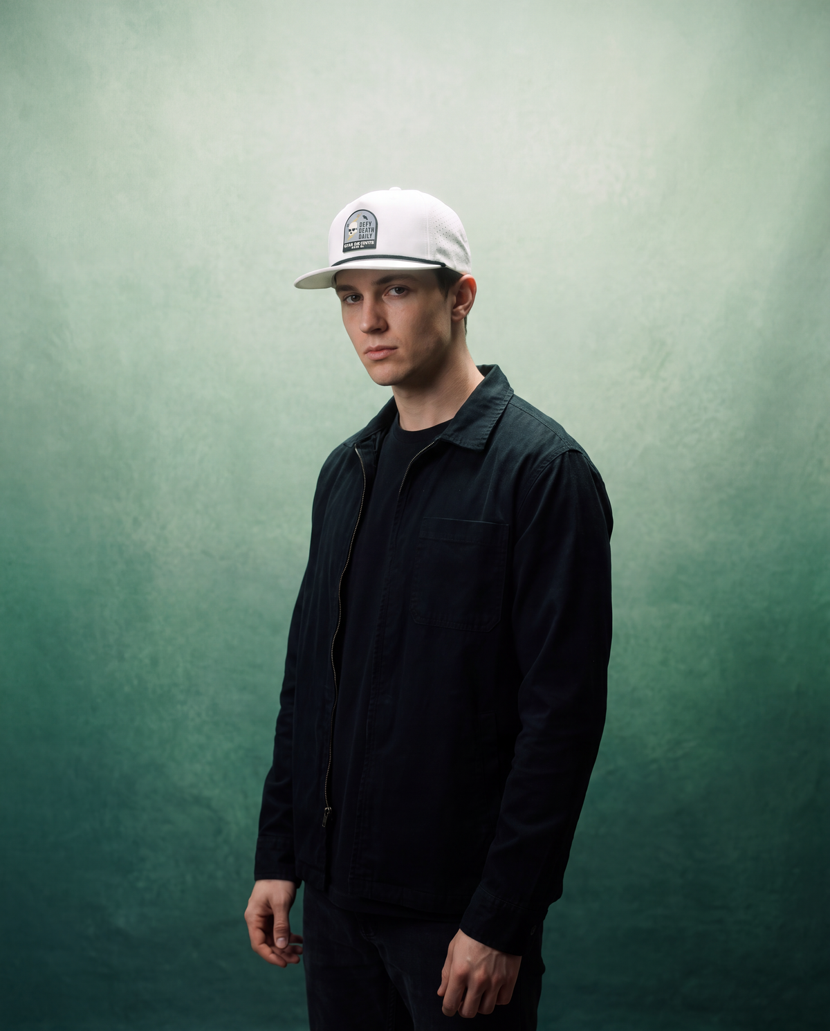 A man wearing a white baseball cap with a black graphic logo, a dark navy zip-up jacket over a black shirt, and dark pants, standing against a textured green studio backdrop.