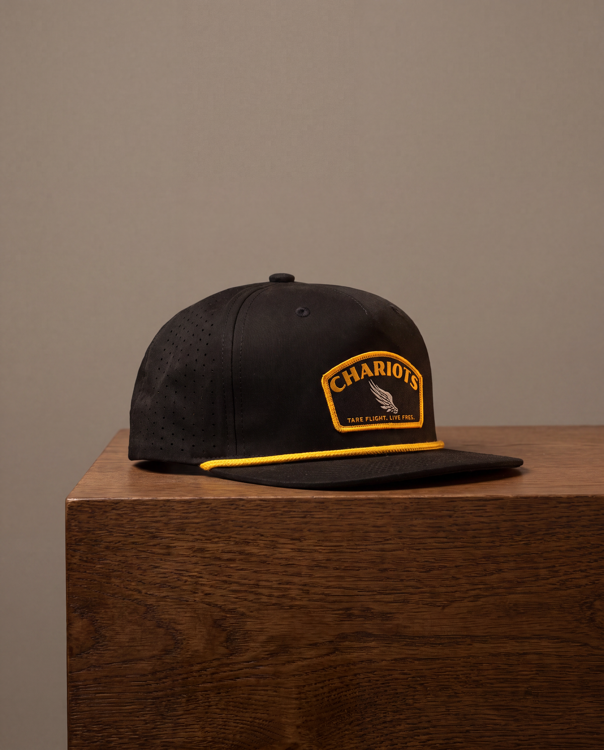 A black flat-brim cap with a yellow embroidered patch reading 'CHARIOTS' and a yellow piping accent along the brim, displayed on a wooden block against a neutral beige background.