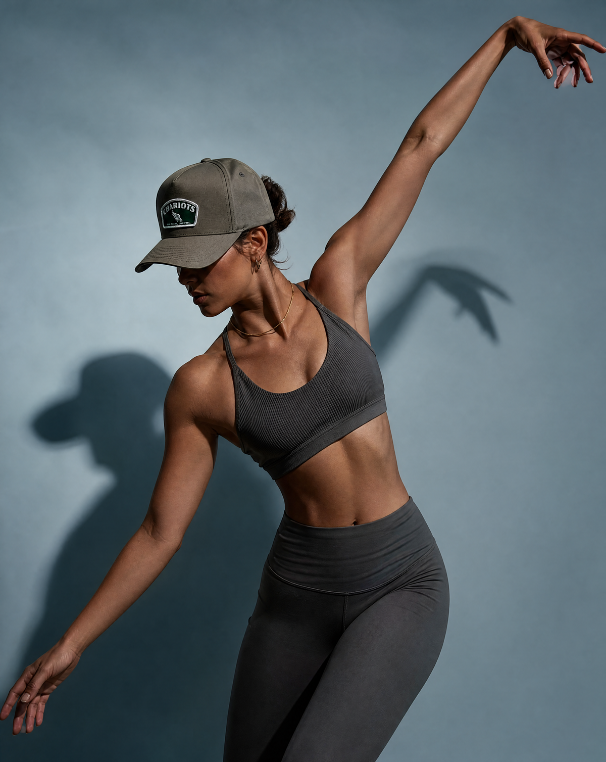 A woman in a dark gray sports bra and matching high-waisted leggings poses dynamically against a slate blue-gray background, wearing a gray baseball cap with a visible 'CHARIOTS' logo patch.