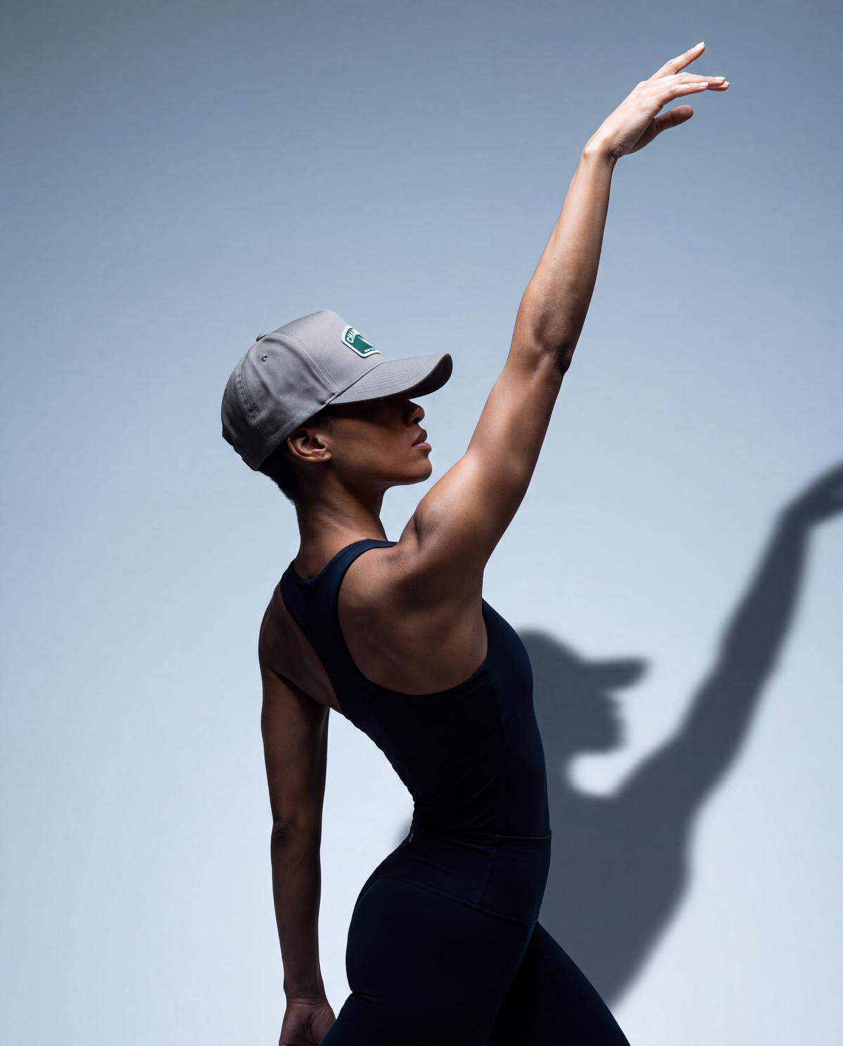 A woman in a black sleeveless bodysuit and gray baseball cap poses with one arm raised against a light gray studio background, casting a long shadow.