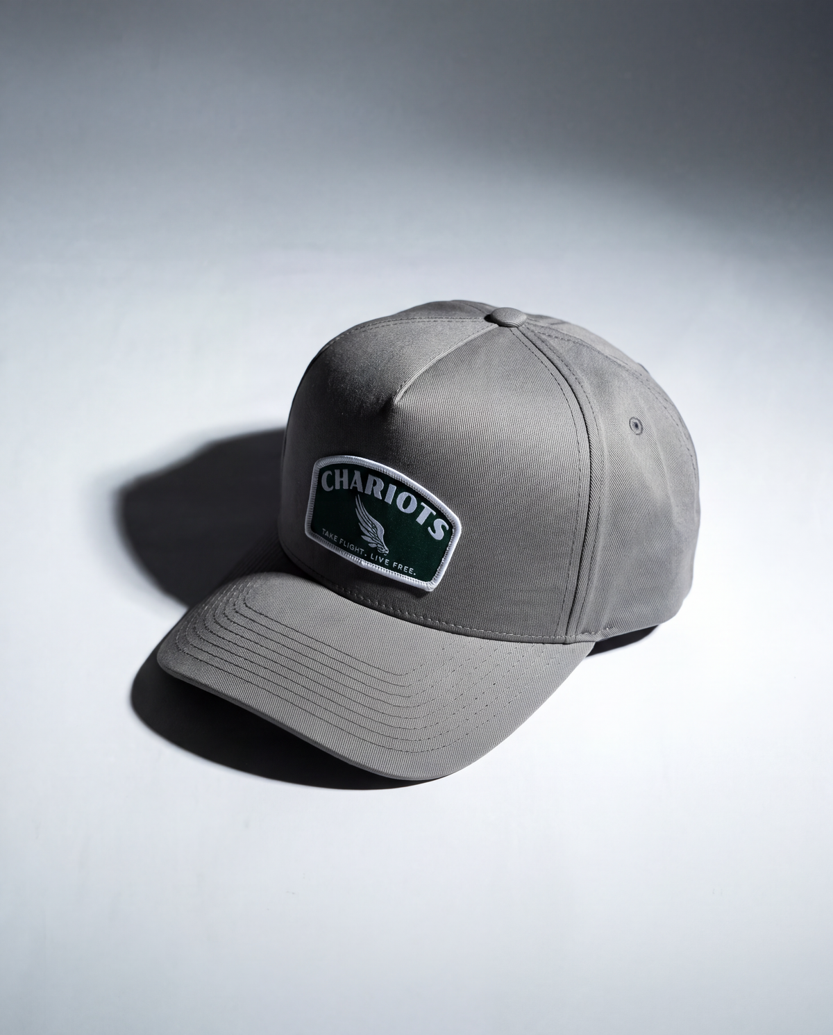 A gray baseball cap with a curved brim, featuring a front patch that reads 'CHARIOTS' with a winged logo and 'TAKE FLIGHT. LIVE FREE' below it. The cap is photographed on a light gray studio background with a soft shadow.