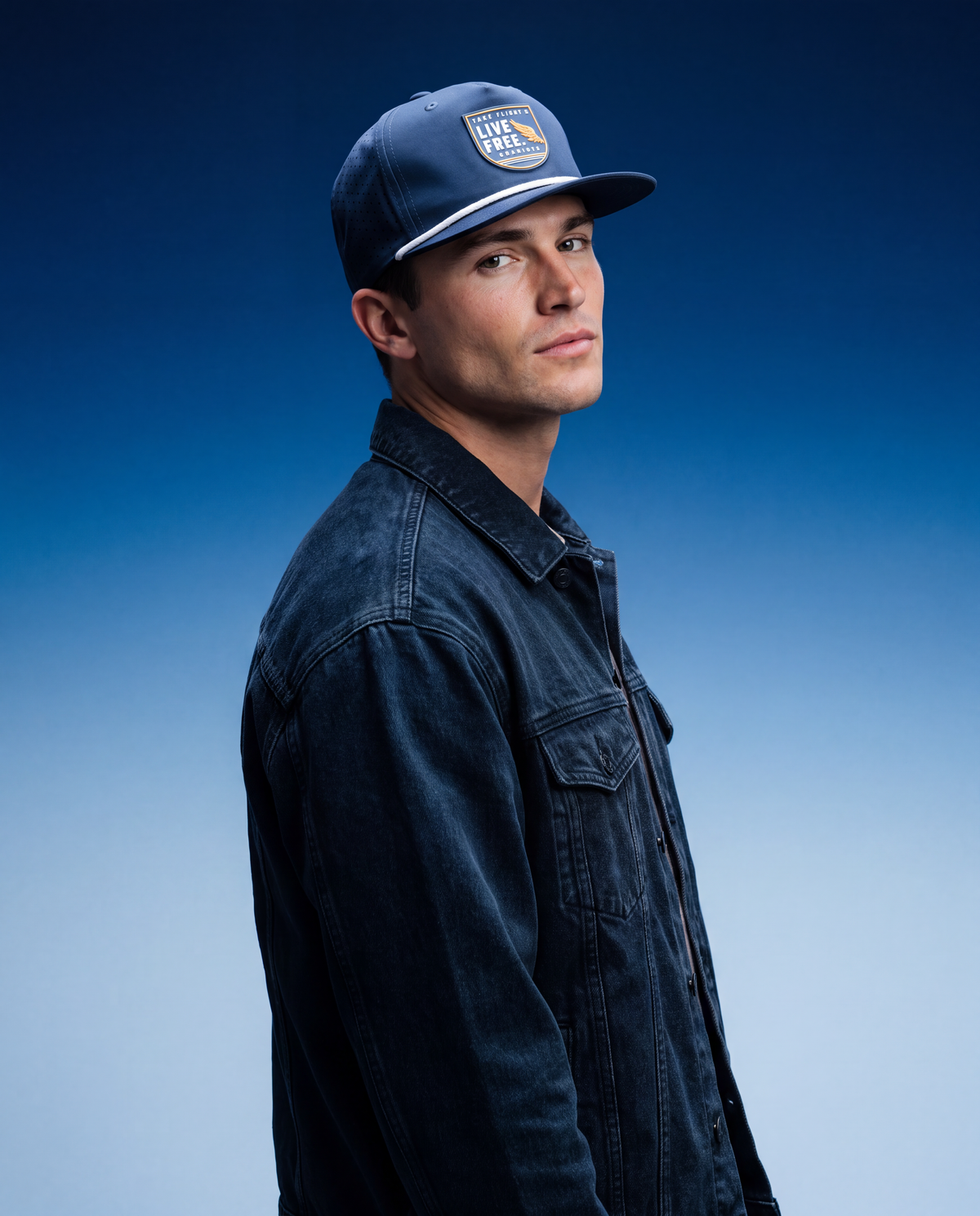 A man in profile wearing a dark blue denim shirt and a matching blue baseball cap with a 'LIVE FREE' logo patch, set against a blue gradient studio background.