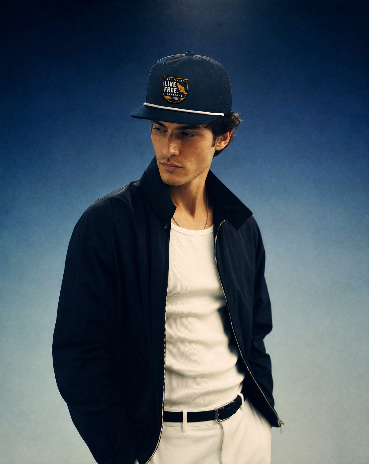 A man wearing a dark navy blue baseball cap with a 'LIVE FREE' patch, a dark navy jacket over a white t-shirt, and white pants, posed against a blue gradient studio background.