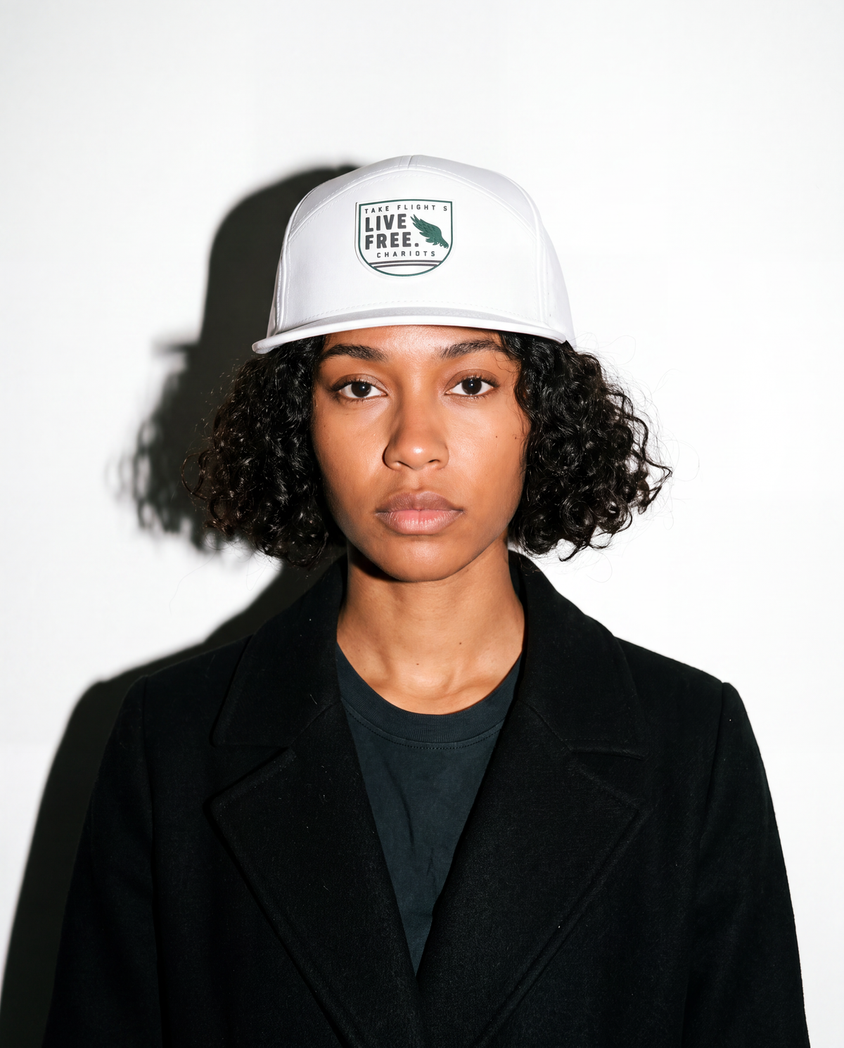 A person with short curly dark hair wearing a white baseball cap with a green and black 'LIVE FREE.