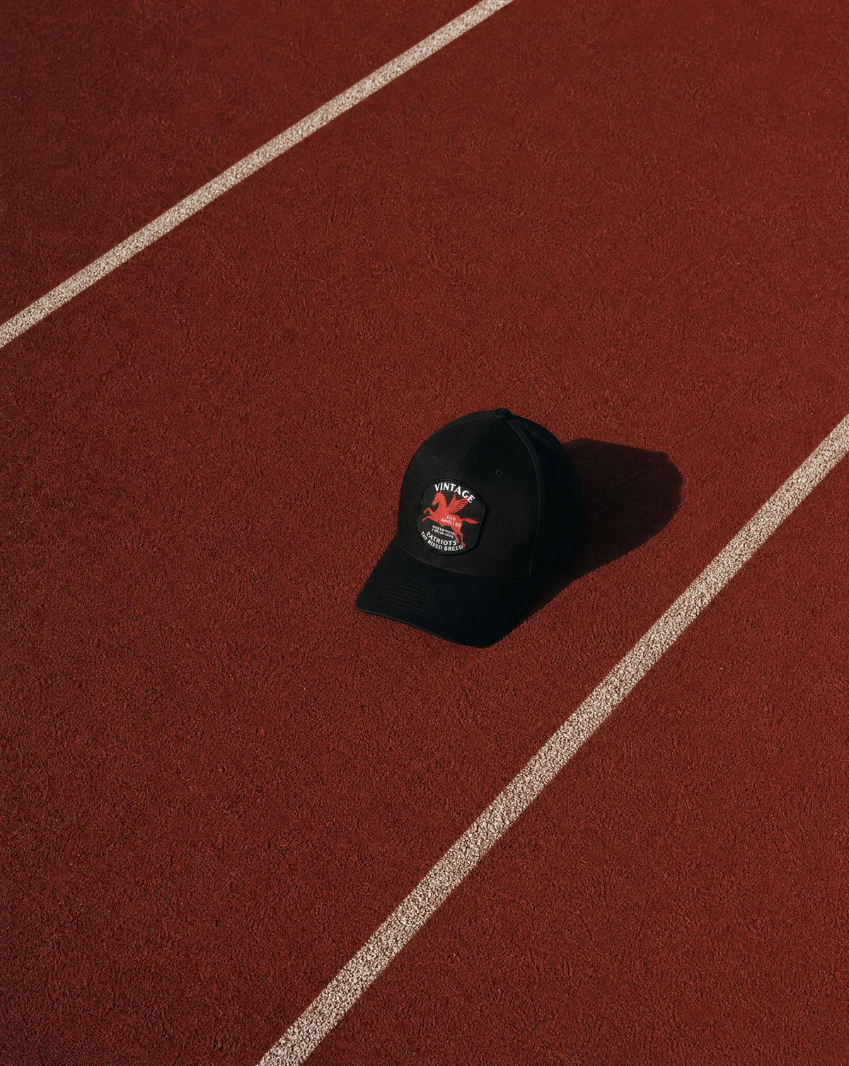 A black baseball cap with a red and white embroidered logo sits on a red running track with white lane lines.