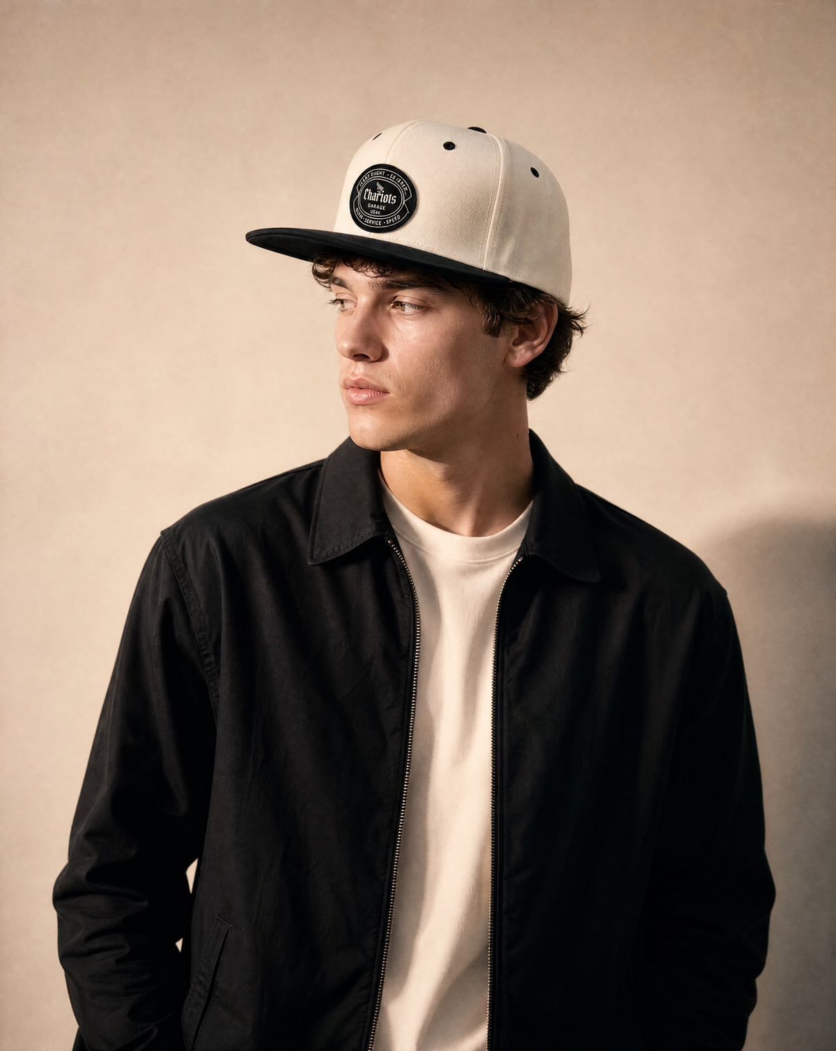 A young man wearing a cream-colored snapback cap with a black brim and a circular black logo patch, styled with a black jacket over a cream t-shirt, set against a warm beige textured background.