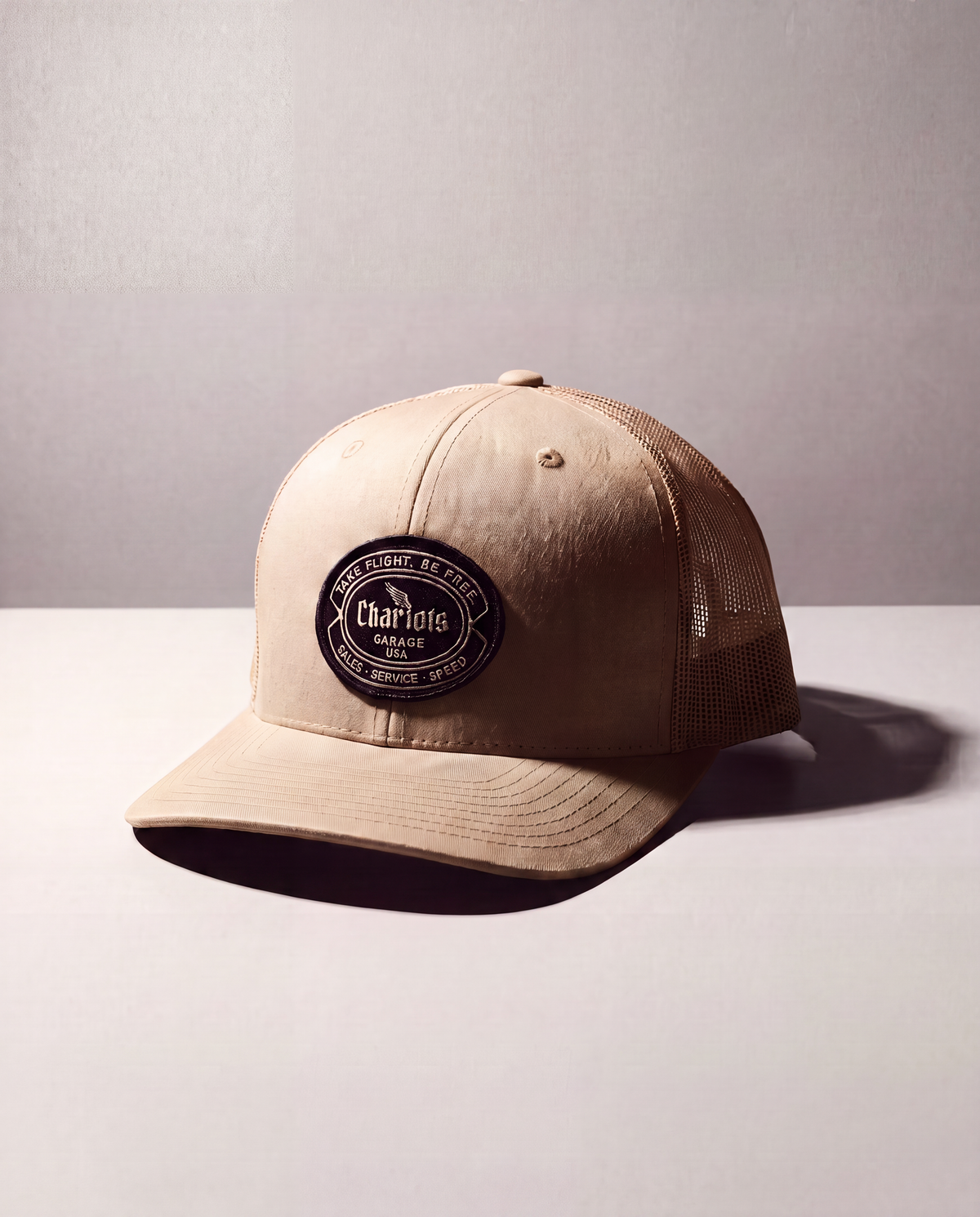 A beige trucker cap with a black circular patch on the front that reads 'Chariots Garage', set against a neutral gray background.