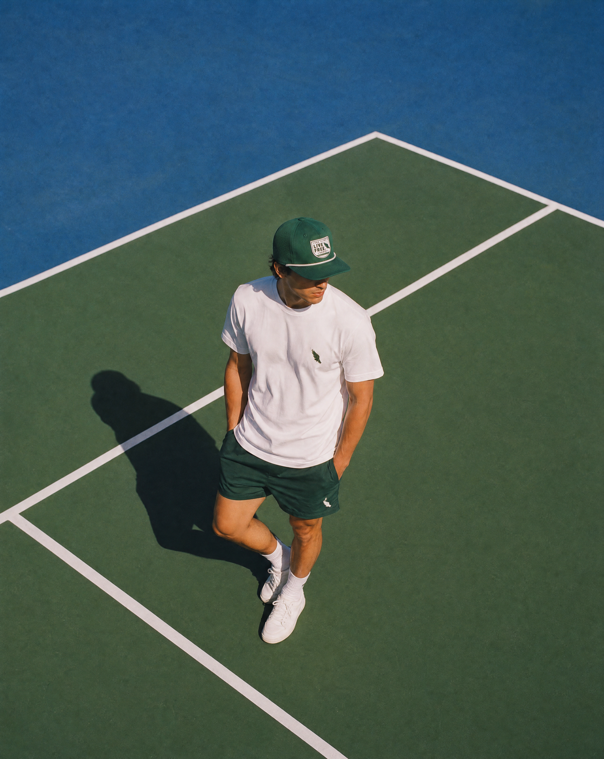 A man wearing a green cap, white t-shirt, dark green shorts, and white sneakers stands on a green tennis court with blue lines under bright sunlight.