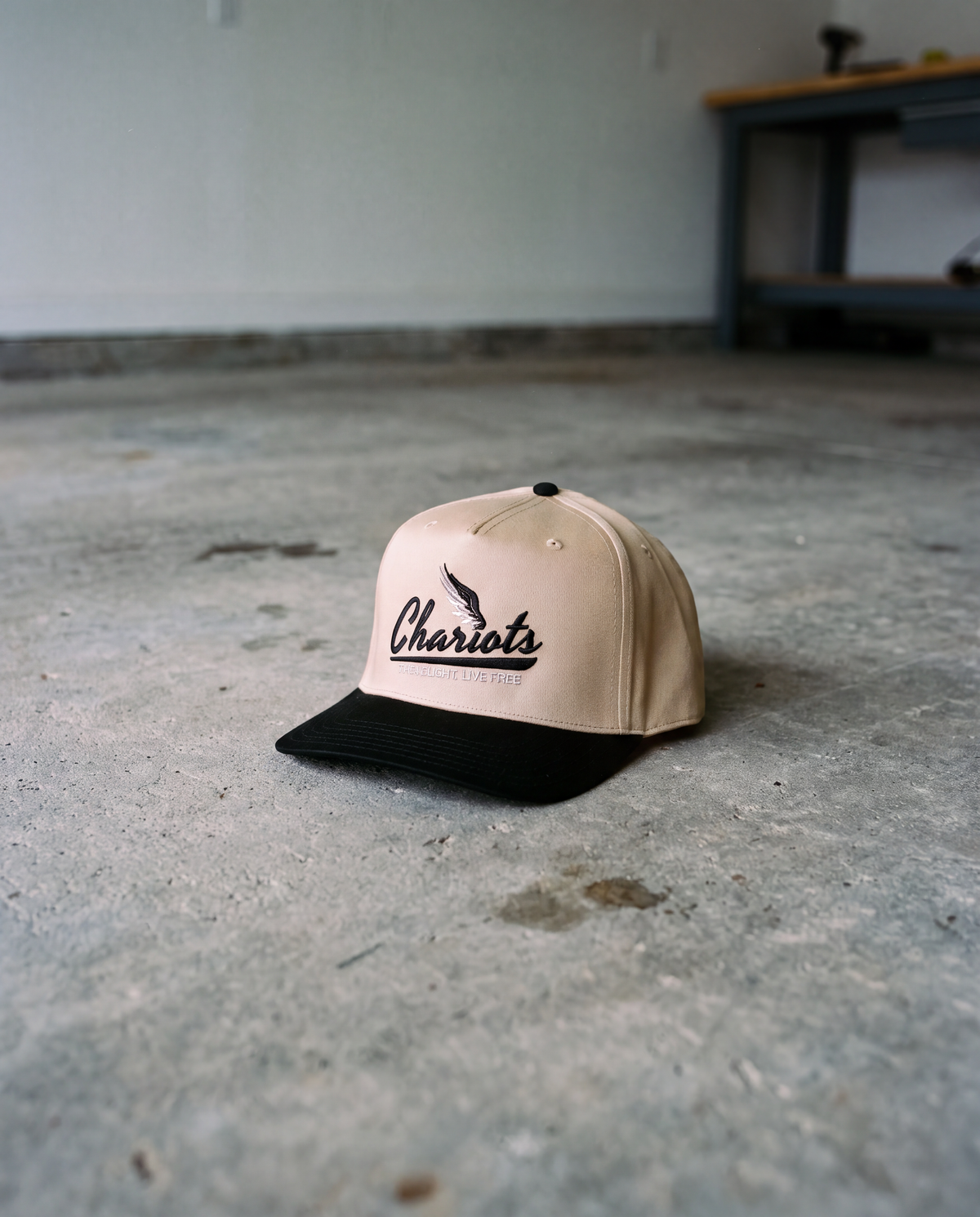 A beige and black snapback cap with 'Chariots' embroidered on the front, resting on a concrete floor in a garage-like setting.