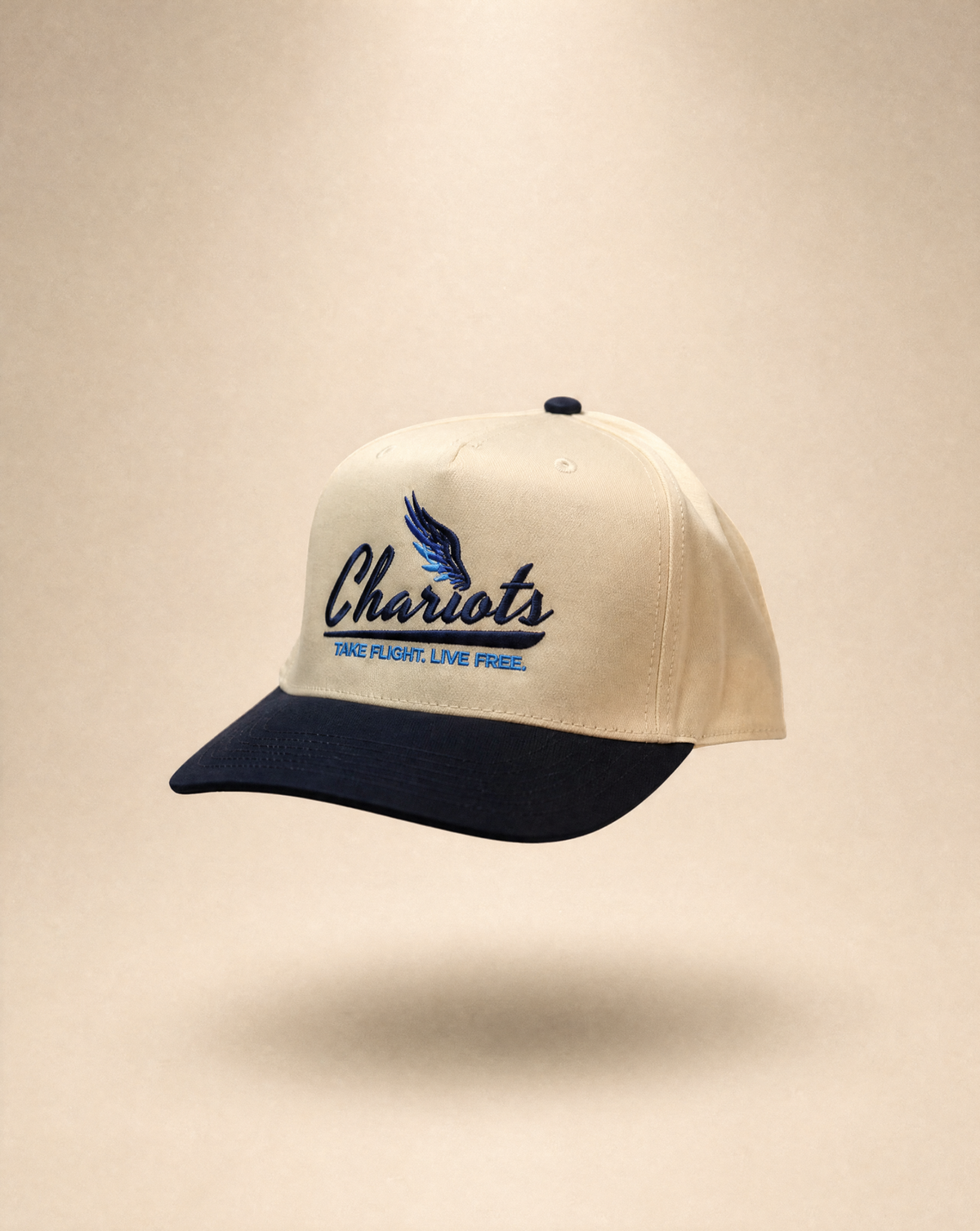 A baseball cap with an off-white crown and a dark navy blue visor, featuring a navy blue embroidered logo that reads 'Chariots' with a wing graphic and the text 'TAKE FLIGHT, LIVE FREE.' on the front, set against a warm beige background.