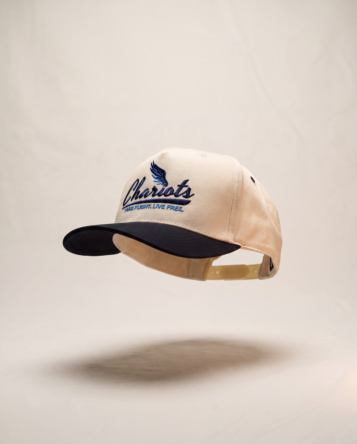A baseball cap with a cream-colored crown and a dark navy blue visor, featuring 'Chariots' embroidered in navy blue on the front with a wing logo. The cap is shown floating against a neutral beige background with a soft shadow underneath.