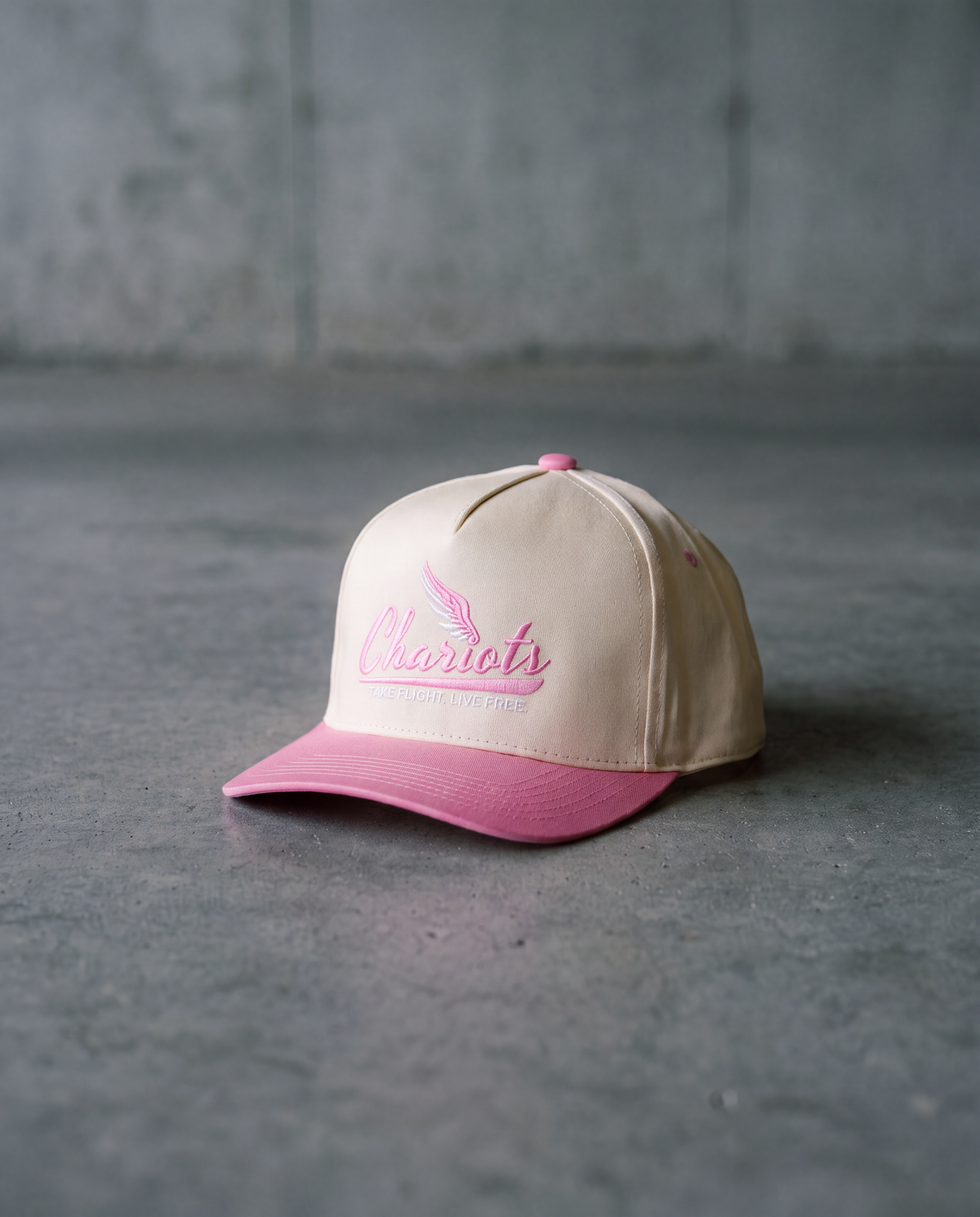A beige baseball cap with a pink visor and pink embroidered logo reading 'Majestic' on the front, photographed on a gray concrete surface against a gray wall.