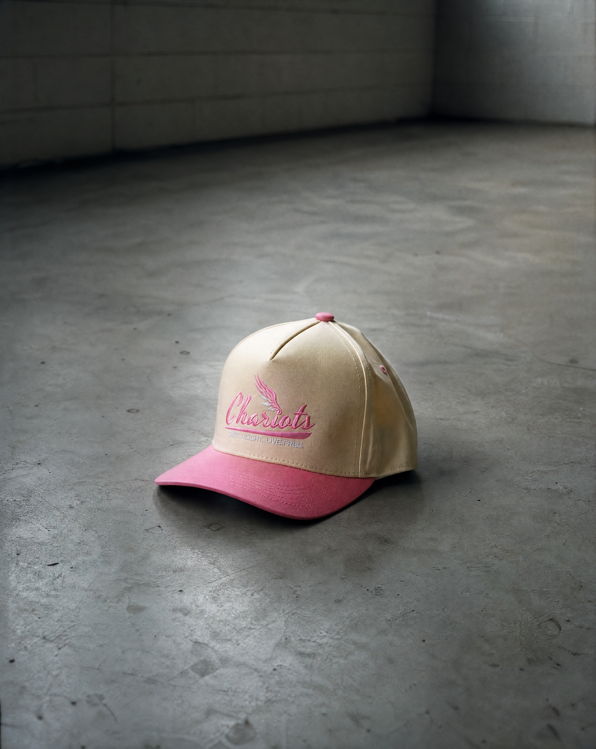 A beige baseball cap with a pink brim and pink embroidered script reading 'ChARIOTS' on the front, placed on a gray concrete floor against a gray brick wall.