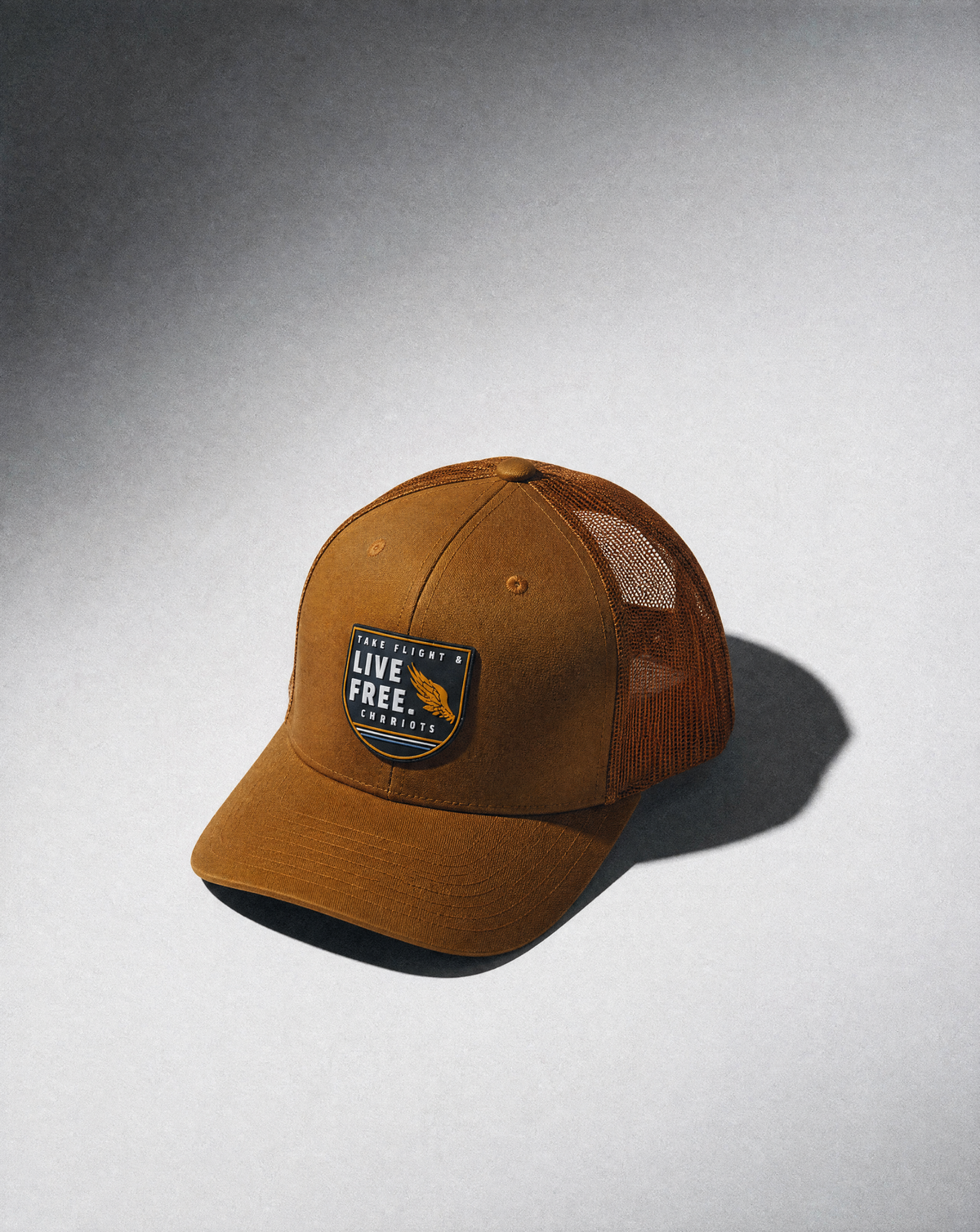 A brown trucker-style cap with a curved brim, featuring a front patch that reads 'TAKE FLIGHT & LIVE FREE' with a bird graphic, set against a gray gradient background with a soft shadow.