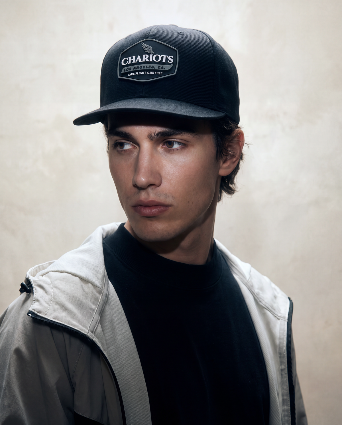 A man wearing a black baseball cap with a white 'CHARIOTS' logo patch, a black t-shirt, and a light gray zip-up jacket, set against a textured beige background.