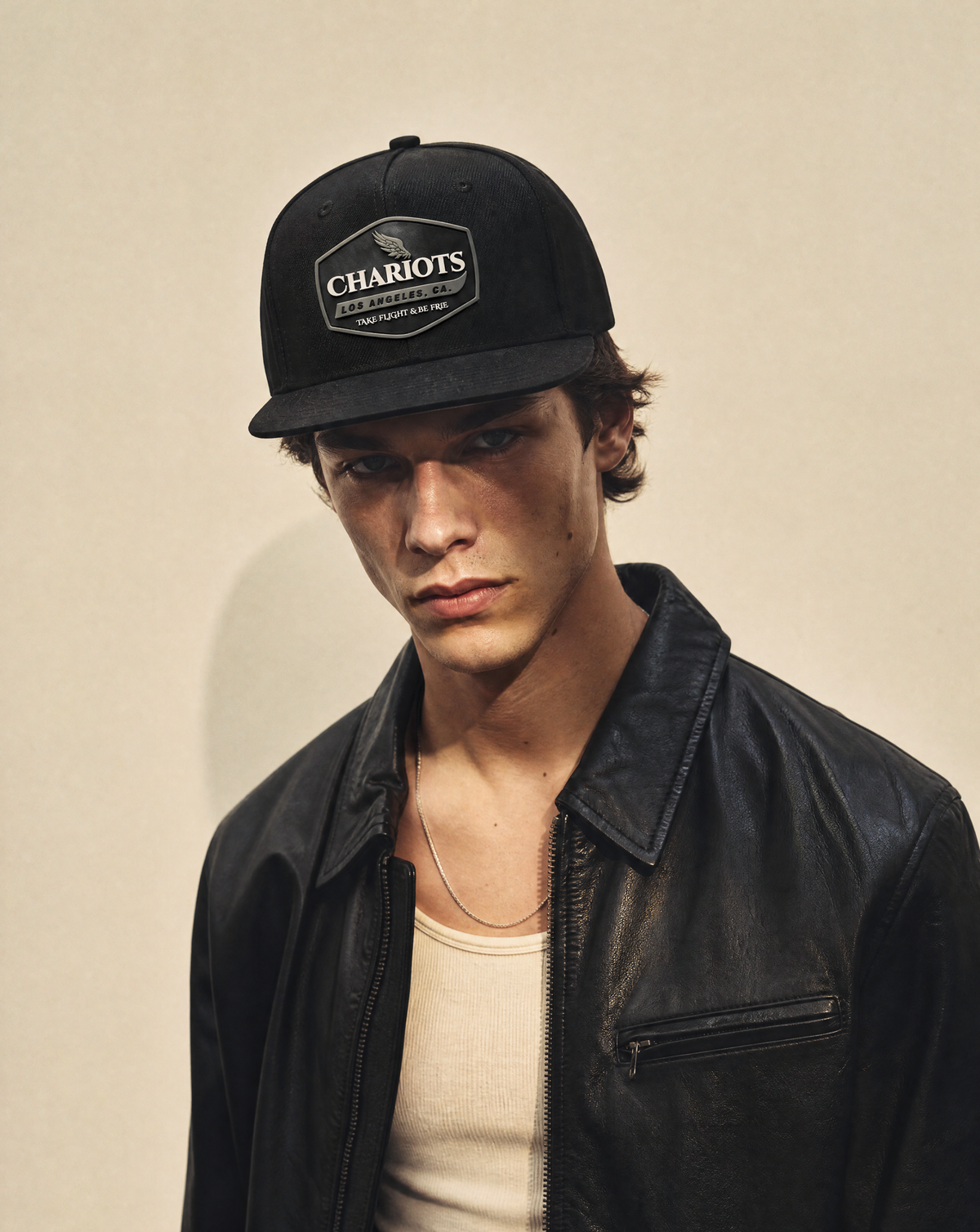A young man wearing a black baseball cap with a 'CHARIOTS' logo patch and a black leather jacket over a cream ribbed t-shirt, photographed against a plain beige background.