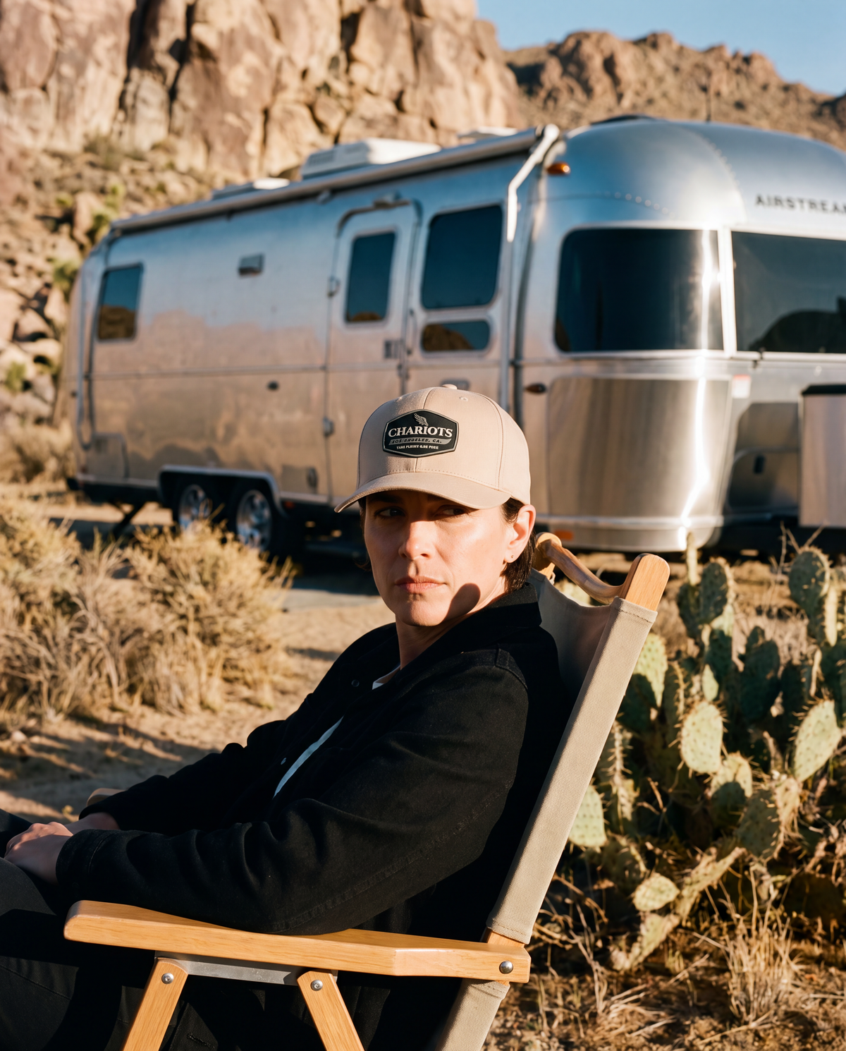 A man wearing a beige cap with a 'Chariots' logo sits in a wooden folding chair in a desert setting, with a silver Airstream trailer in the background.