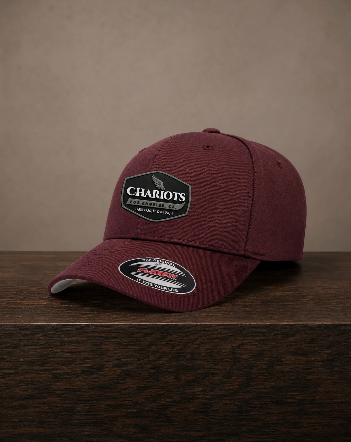 A maroon baseball cap with a black and white Chariots Aviation patch on the front, resting on a dark wooden surface against a textured beige background.