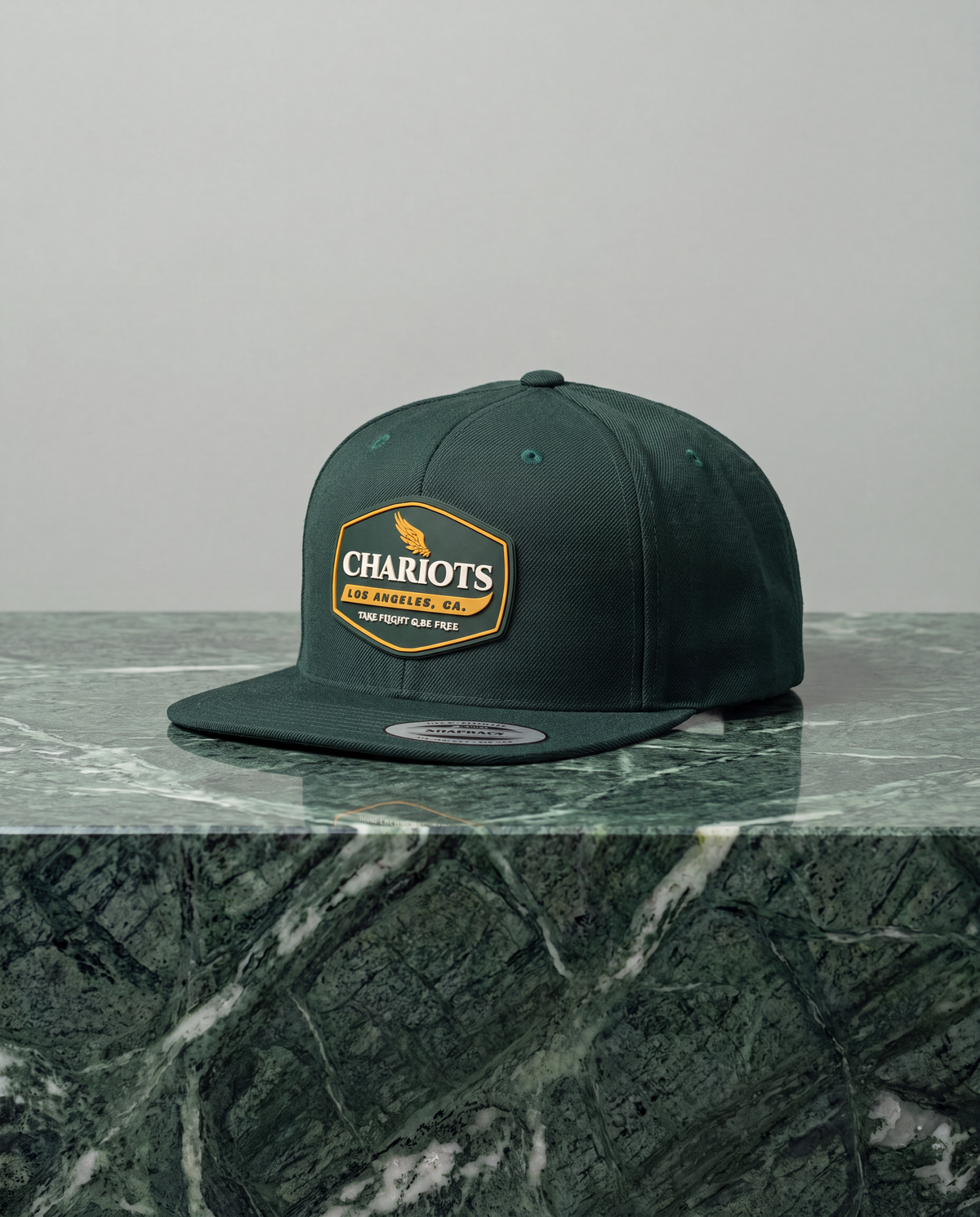 A dark forest green flat-brim cap with a gold and white 'Chariots' patch on the front, displayed on a green marble surface against a light gray background.