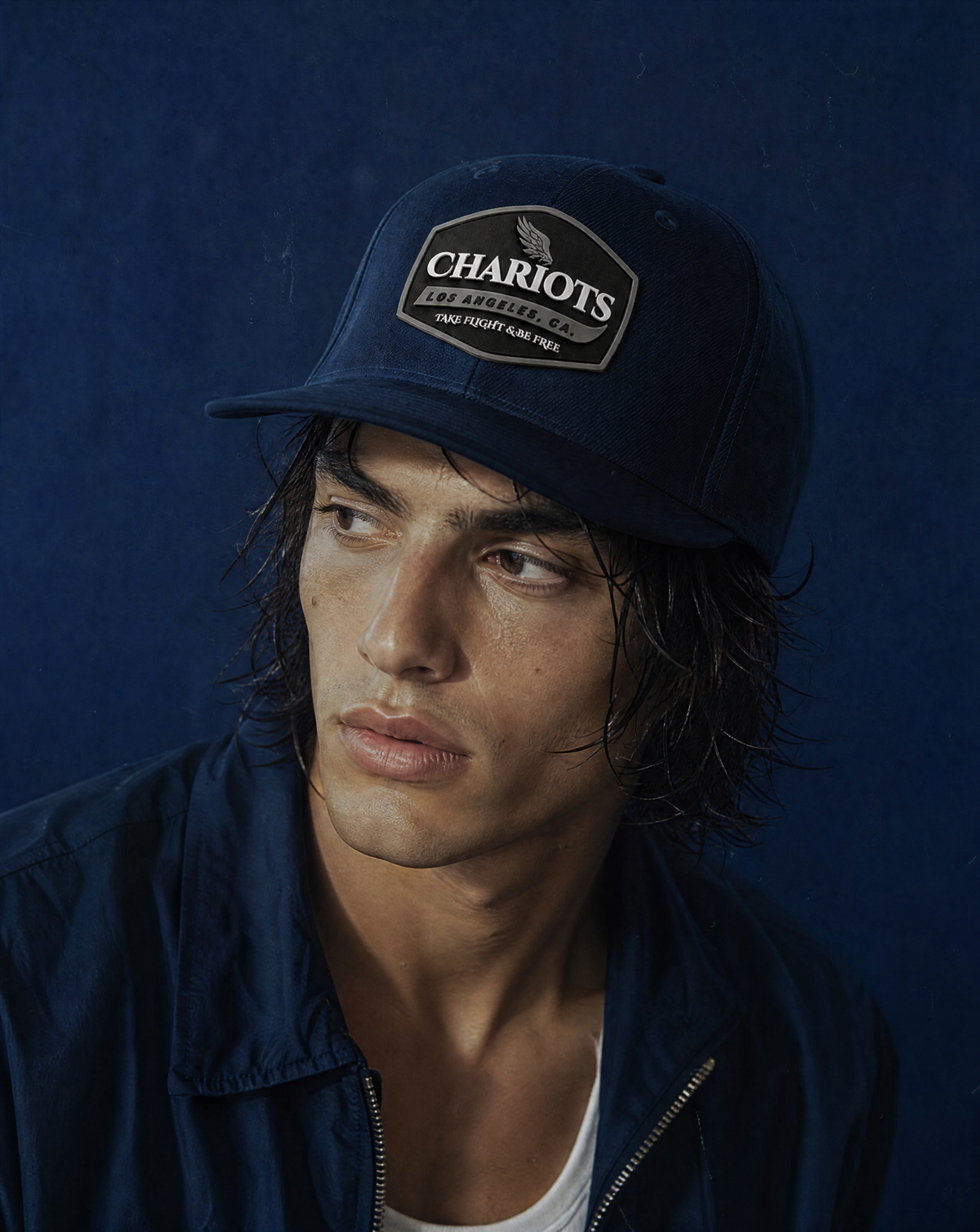 A man wearing a navy blue baseball cap with a 'Chariots' embroidered patch and a matching navy blue jacket, set against a dark blue background.