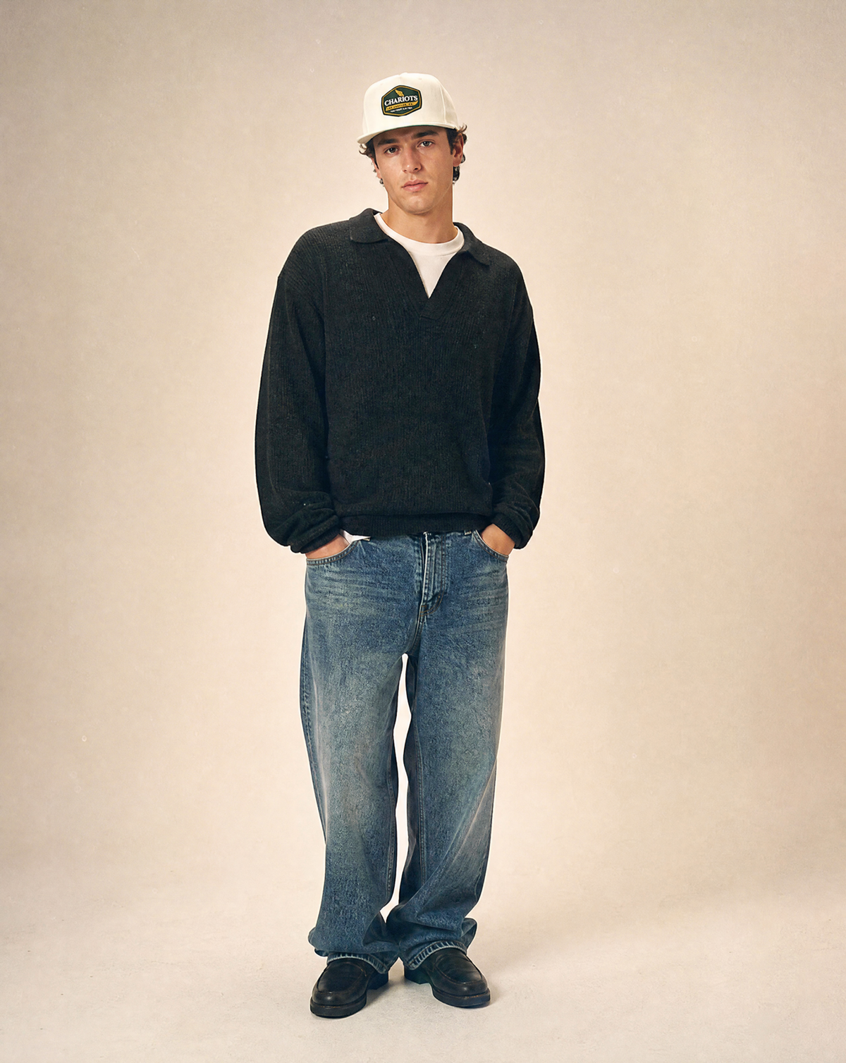 A man stands against a plain beige studio background wearing a white baseball cap with a green logo, a dark charcoal knit polo-style sweater over a white undershirt, loose blue jeans, and black shoes.