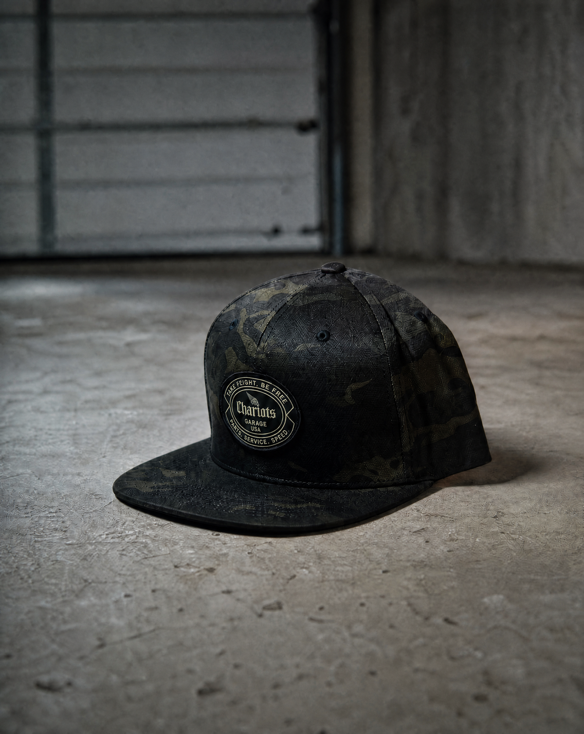 A dark camouflage flat-brim cap with a circular embroidered 'Chariots' logo patch on the front, resting on a concrete floor in a dimly lit industrial setting.