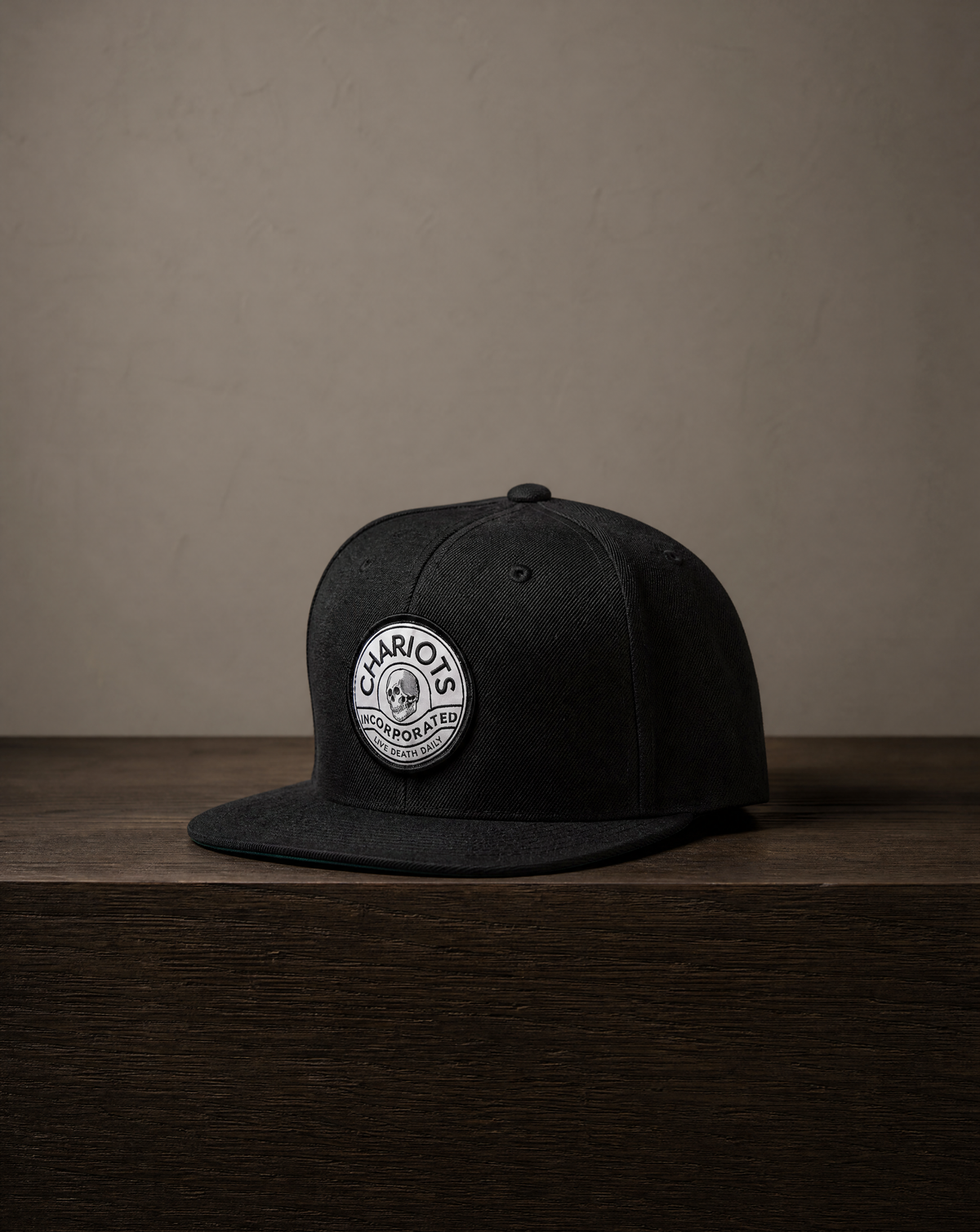 A black flat-brim snapback cap with a circular white embroidered patch on the front, displayed on a dark wooden surface against a textured beige wall.
