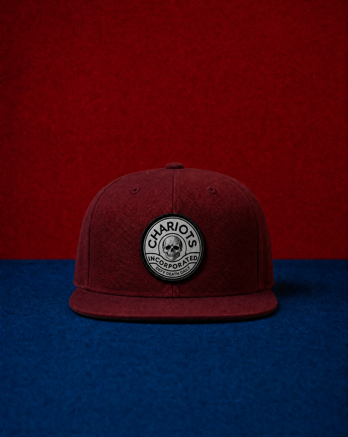 A dark red flat-brim cap with a circular embroidered patch on the front that reads 'CHARIOTS INCORPORATED' around a skull logo, set against a split red and blue background.