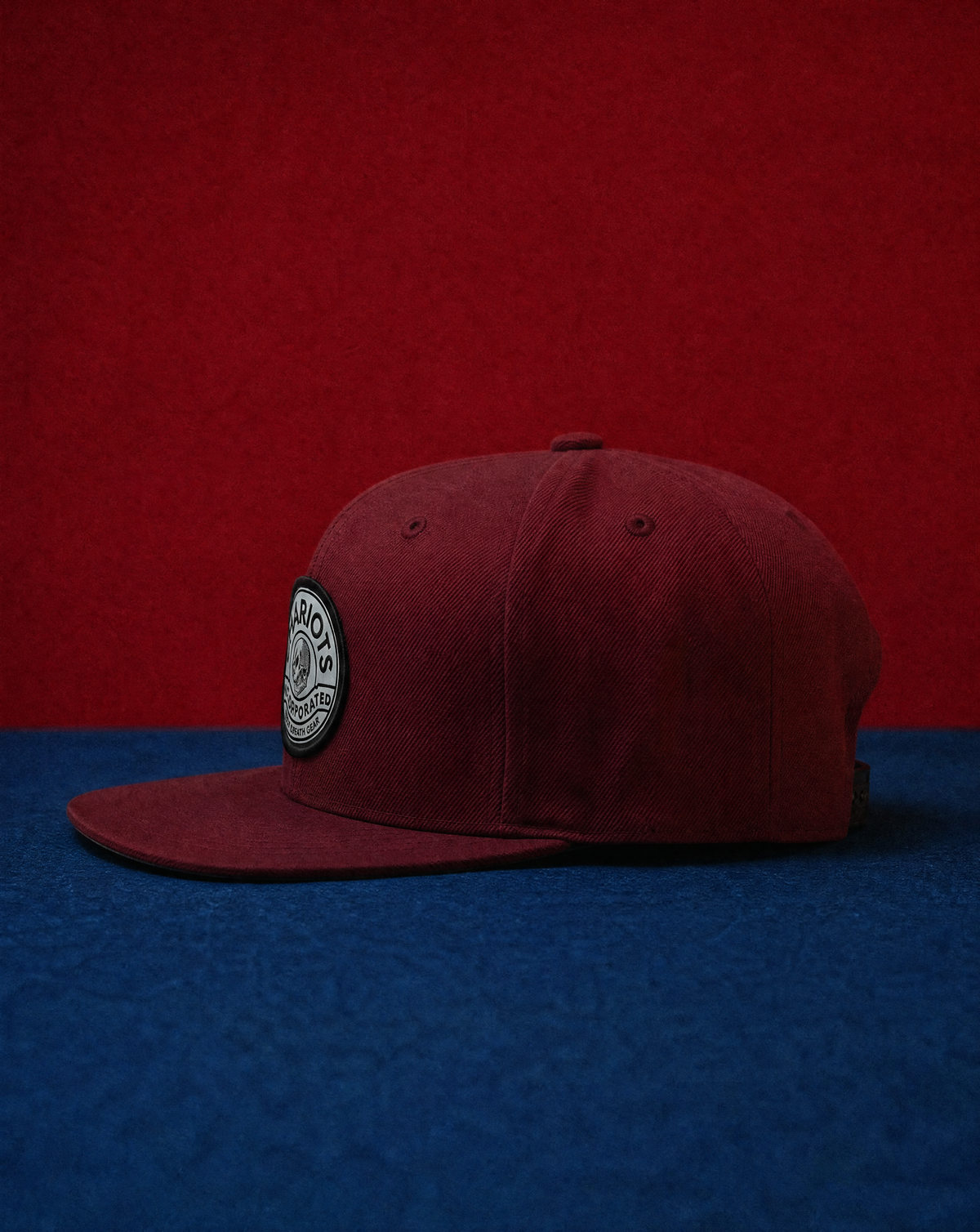 A structured flat-brim cap in deep burgundy, featuring a circular black and white embroidered patch on the side, set against a split red and blue textured background.