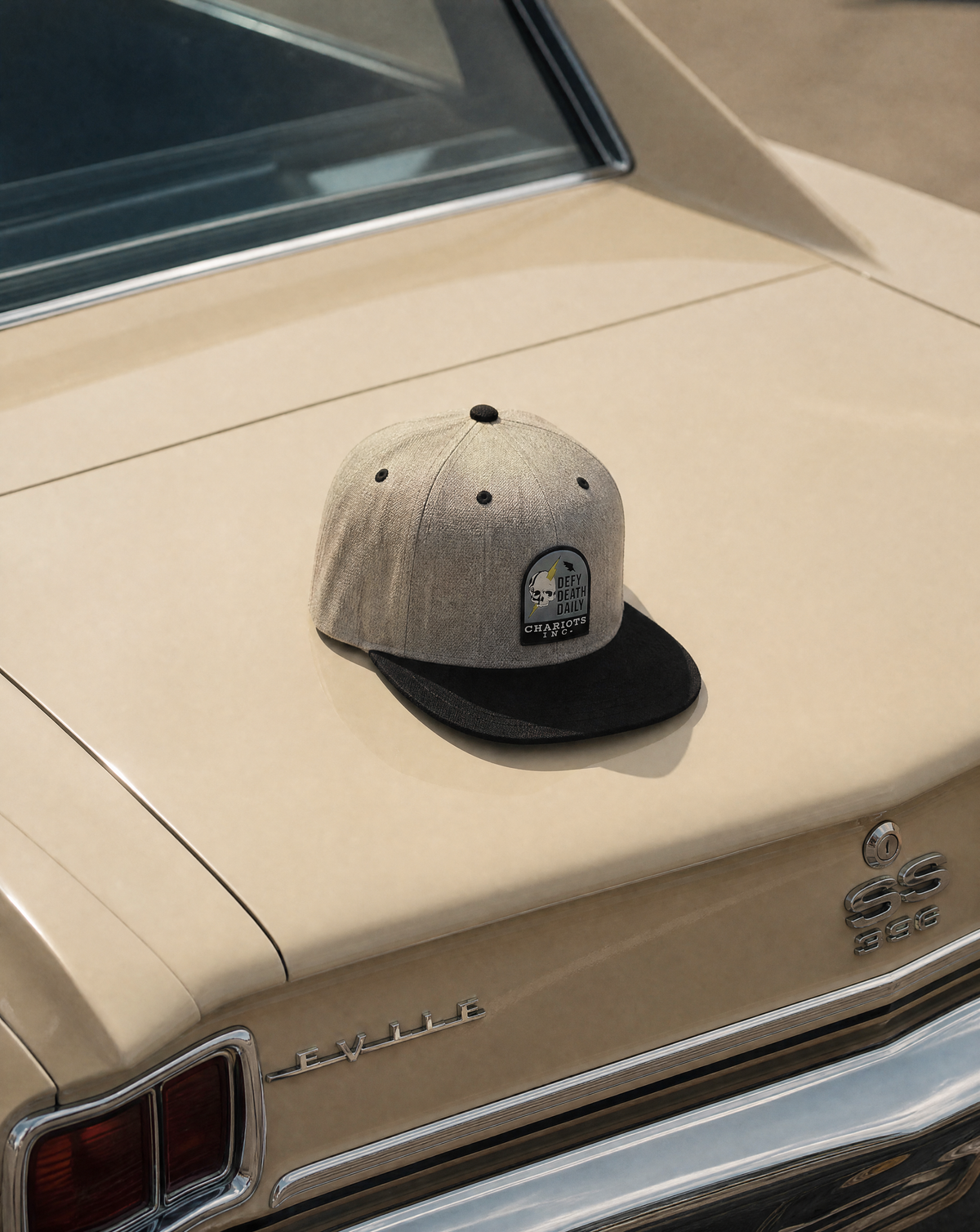 A light gray heathered flat-brim cap with a black visor sits on the trunk of a beige vintage car. The cap features a small embroidered patch on the front.