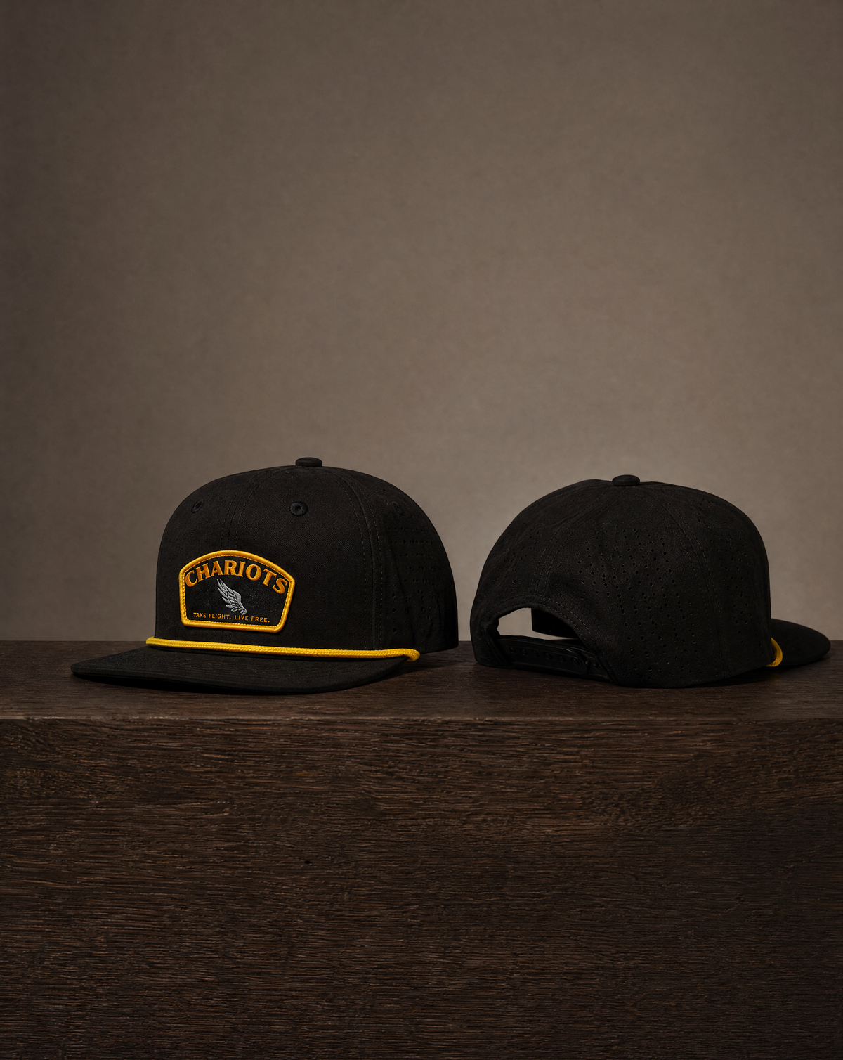 Two black flat-brim snapback caps displayed on a dark wooden surface against a warm taupe background. The left cap shows a front view with a yellow and black 'CHARIOTS' patch and yellow piping, while the right cap shows the back view with an adjustable snapback closure.