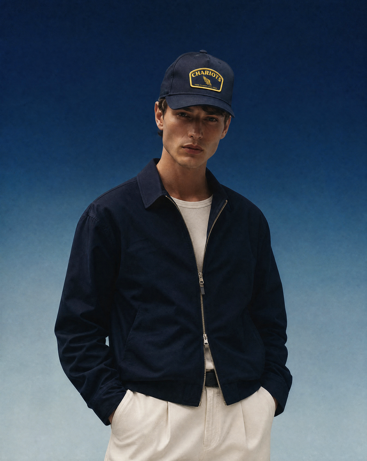 A man wearing a navy blue zip-up jacket, a matching navy blue Chariots baseball cap, a white t-shirt, and off-white trousers, standing against a blue gradient background.