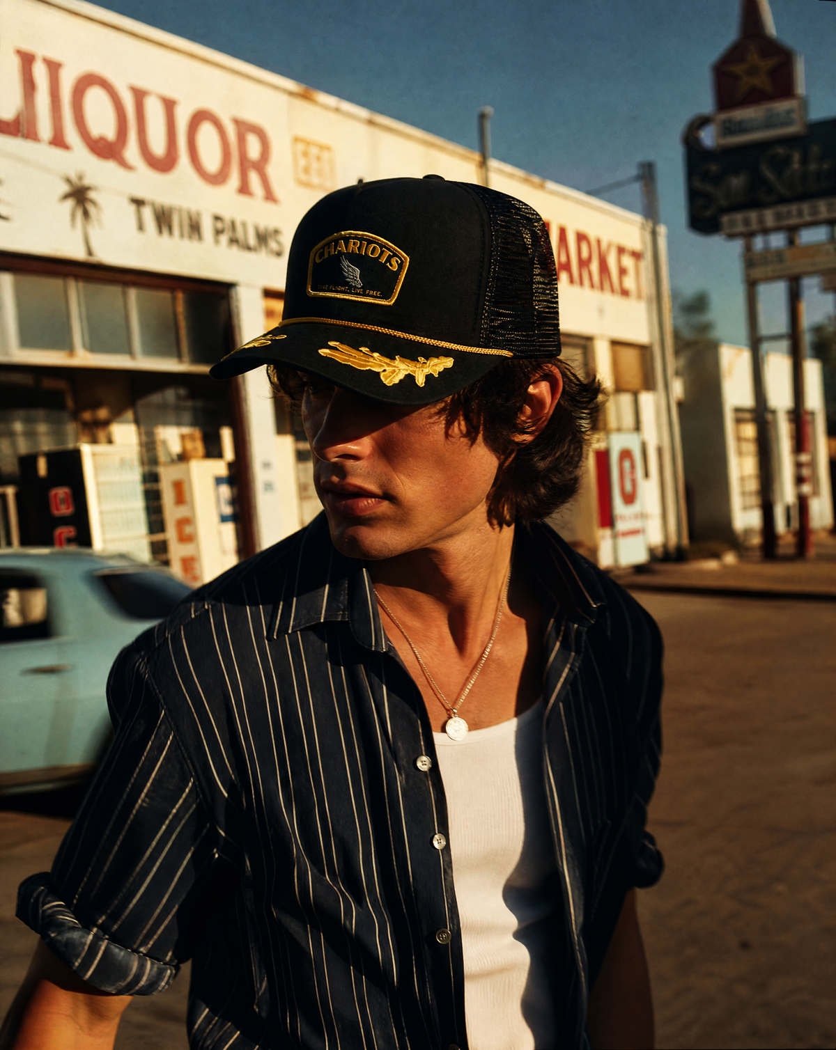 A man wearing a black trucker hat with a gold patch reading 'CHARIOTS' and a gold eagle emblem, paired with a black and white pinstriped button-up shirt over a white undershirt, standing outdoors in front of a vintage gas station building.