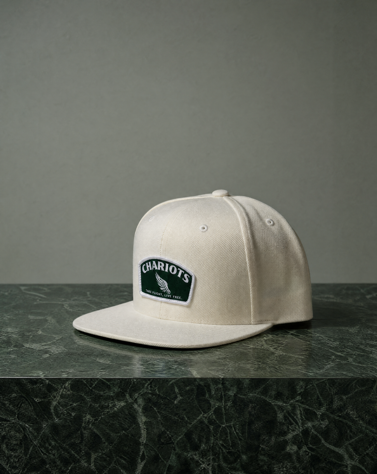 An off-white flat-brim baseball cap with a green and white embroidered 'CHARIOTS' patch on the front, resting on a dark green marble surface against a gray background.