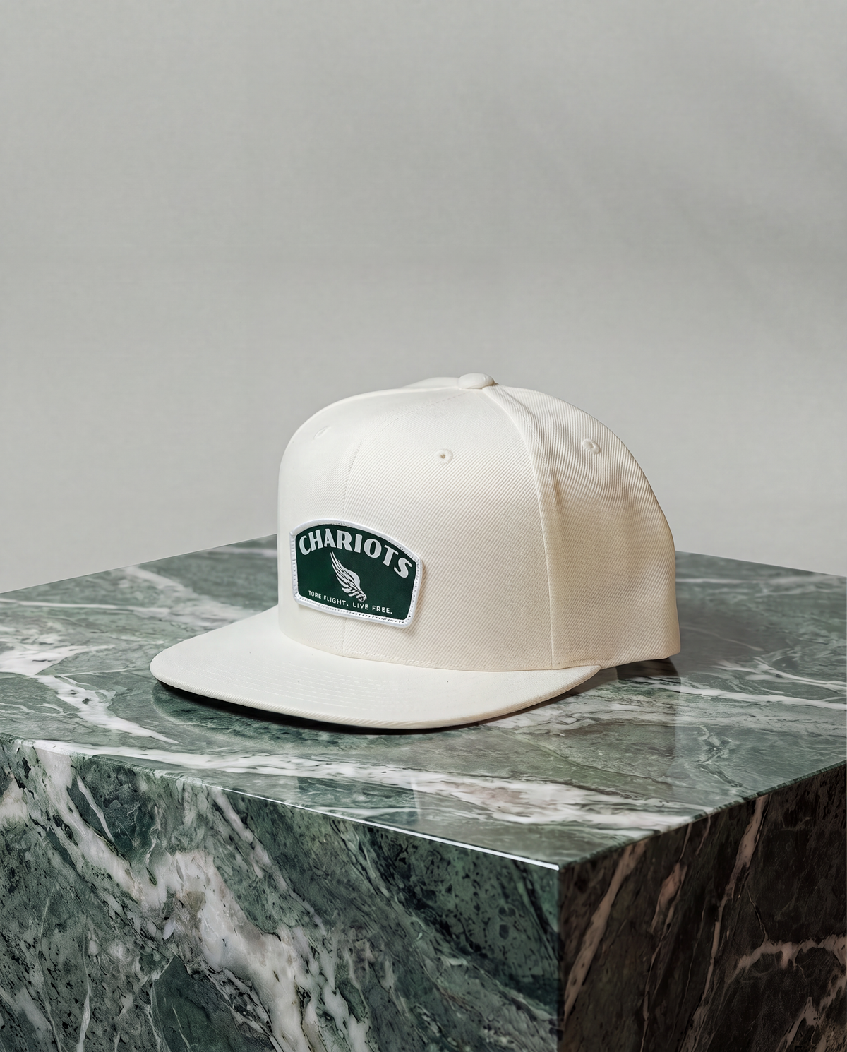 An off-white flat-brim baseball cap with a green and white 'CHARIOTS' patch on the front, displayed on a green marble block against a neutral gray background.