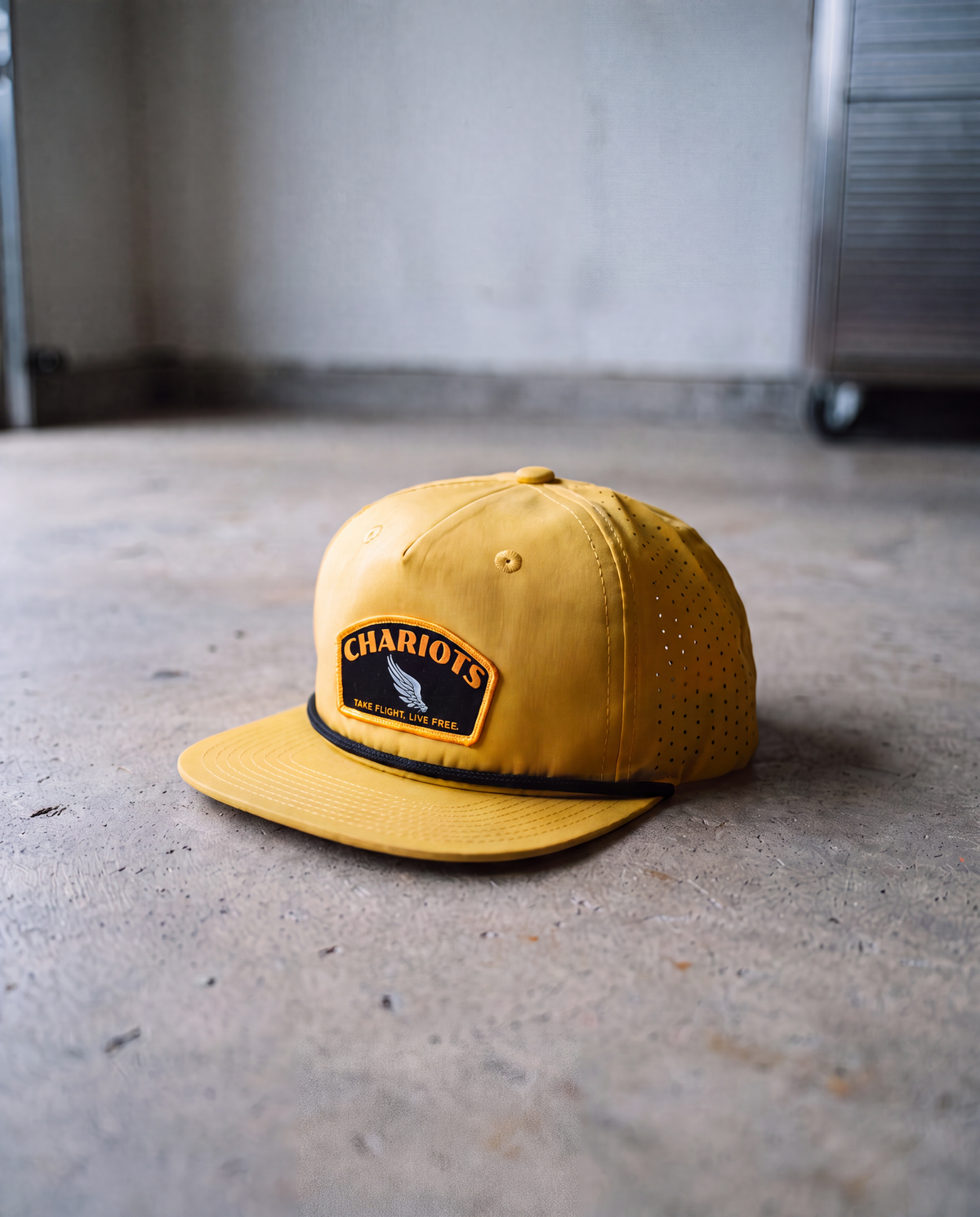 A yellow flat-brim cap with a black band and a front patch reading 'CHARIOTS' and 'TAKE FLIGHT. LIVE FREE', photographed on a concrete floor in an indoor setting.