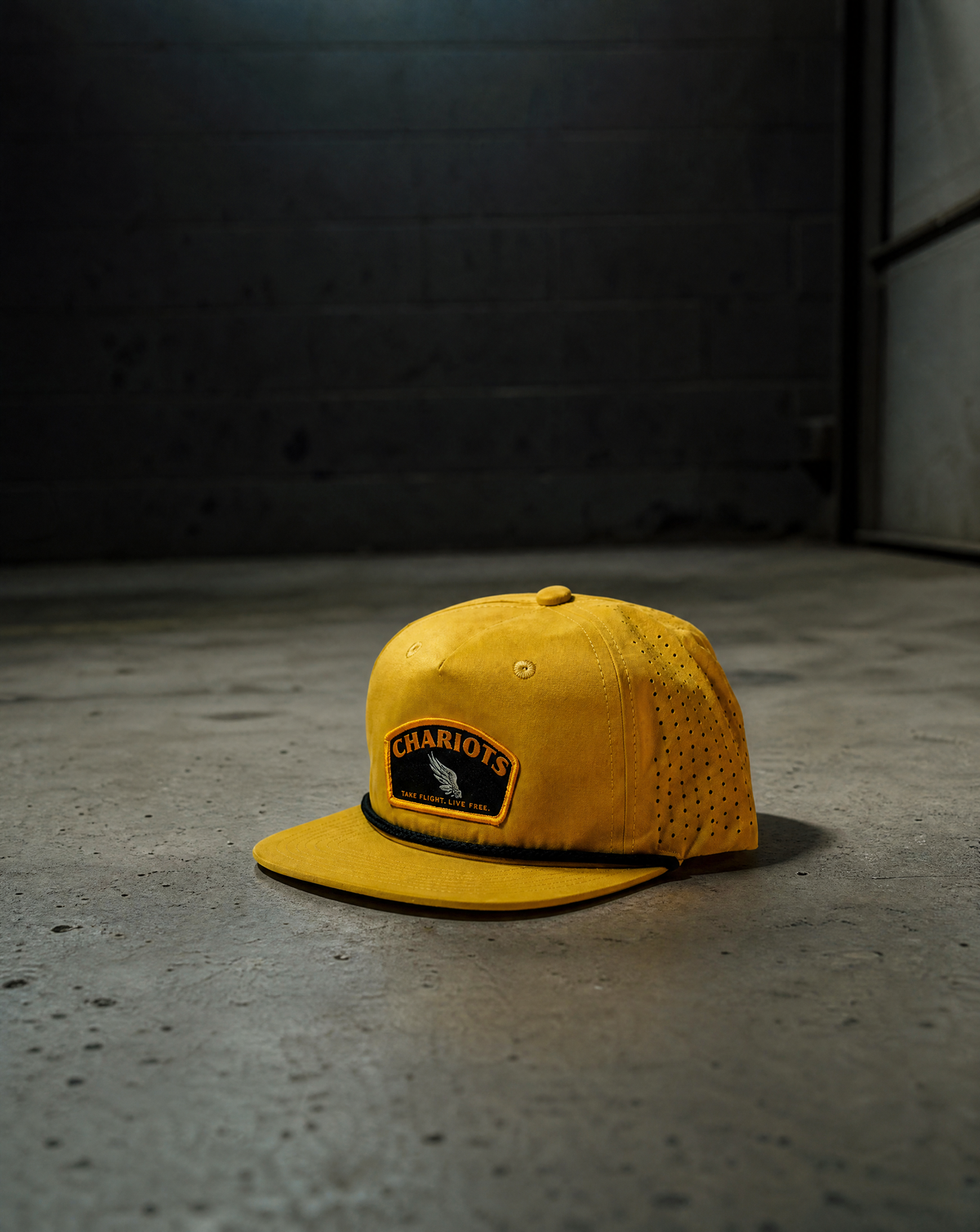 A yellow baseball cap with a black and yellow 'CHARIOTS' patch on the front, resting on a concrete floor against a dark gray brick wall.