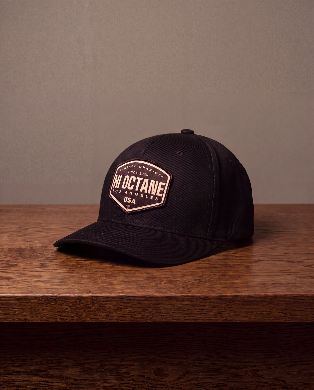 A black baseball cap with a prominent beige and black 'Hi Octane' patch on the front, resting on a dark wooden table against a neutral gray wall.