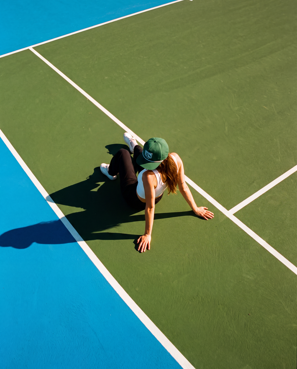 A woman in athletic wear and a green cap poses on a green tennis court with a blue baseline, viewed from a high-angle perspective.