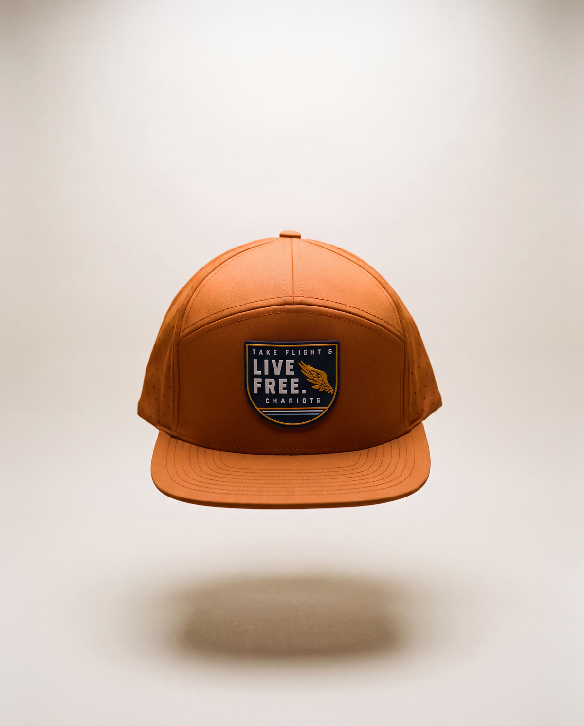 A rust-orange snapback cap with a front patch reading 'TAKE FLIGHT. LIVE FREE, CHARIOTS' and a gold bear logo, shown floating against a light beige background.