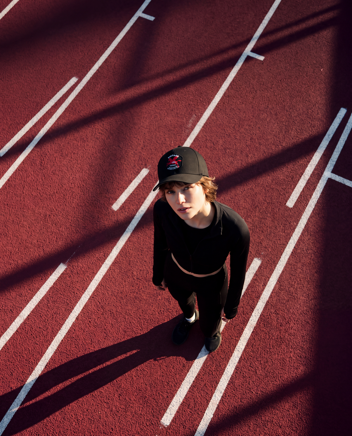 A person standing on a red running track wearing a black long-sleeve crop top, black pants, and a black baseball cap, photographed from a high-angle overhead perspective with strong sunlight and shadows.