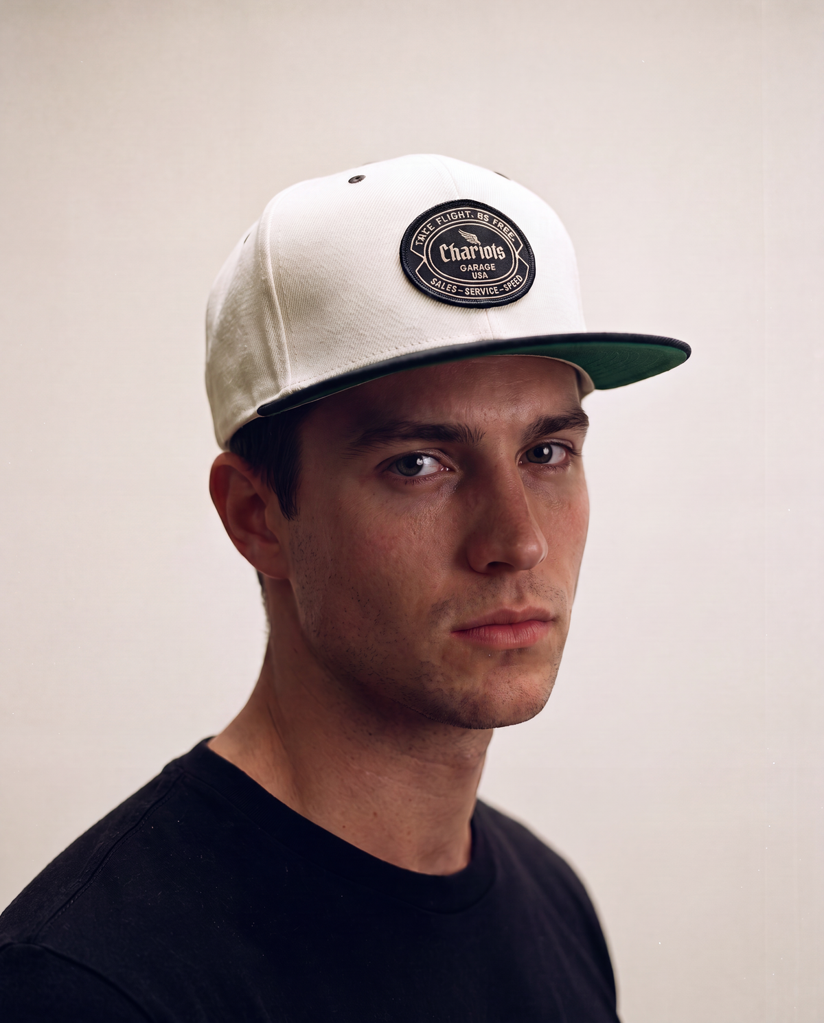 A man wearing a white snapback cap with a dark green brim and a circular black patch on the front that reads 'Chariots'.