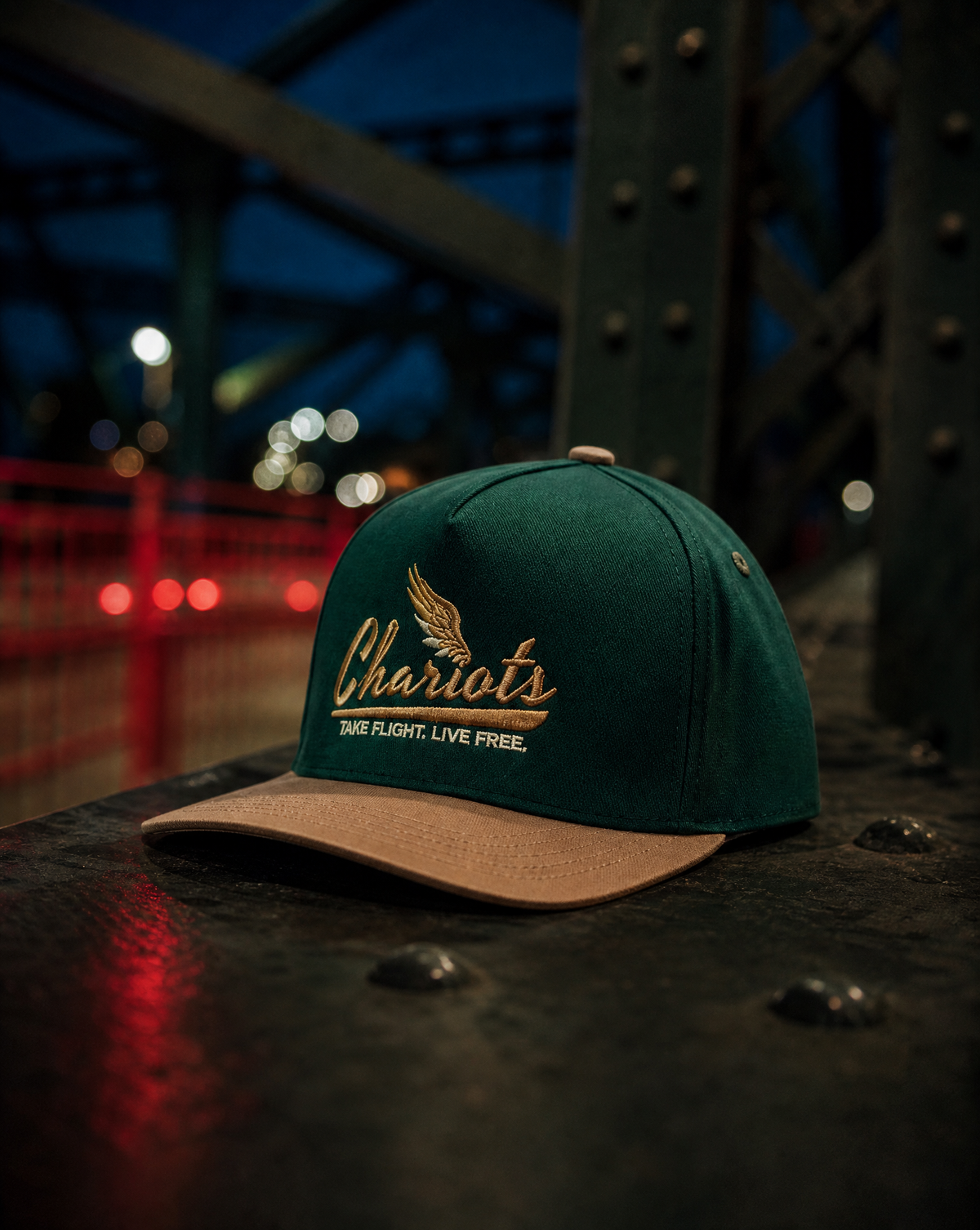A dark green baseball cap with a tan visor, featuring 'Chariots' embroidered in gold on the front, resting on a dark metal surface with a blurred industrial bridge background at dusk.