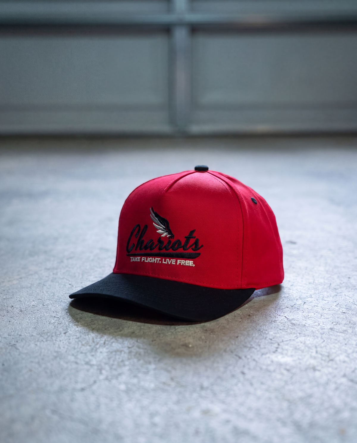 A red and black baseball cap with 'Chariot's' embroidered in black script on the front, resting on a gray concrete floor.