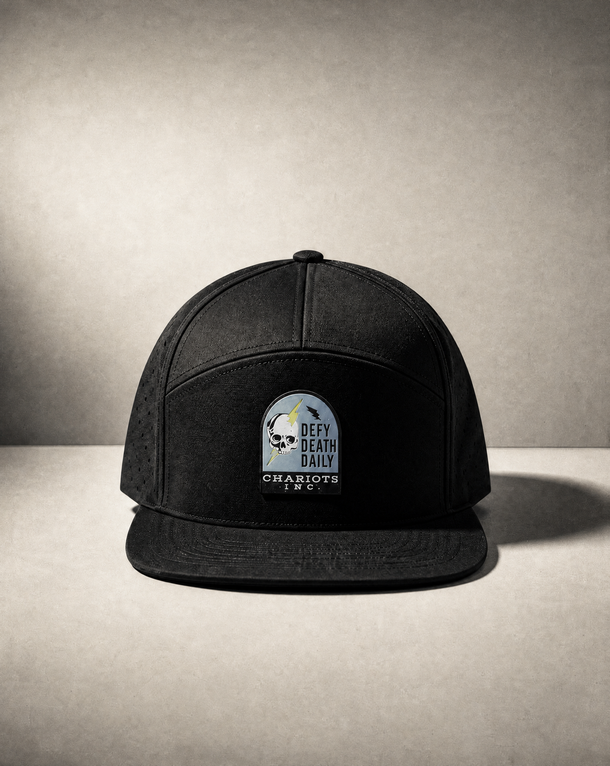 A black flat-brim snapback cap with a circular embroidered patch on the front that reads 'DEATH DAILY' and 'CHARIOTS inC', photographed on a textured gray studio background.